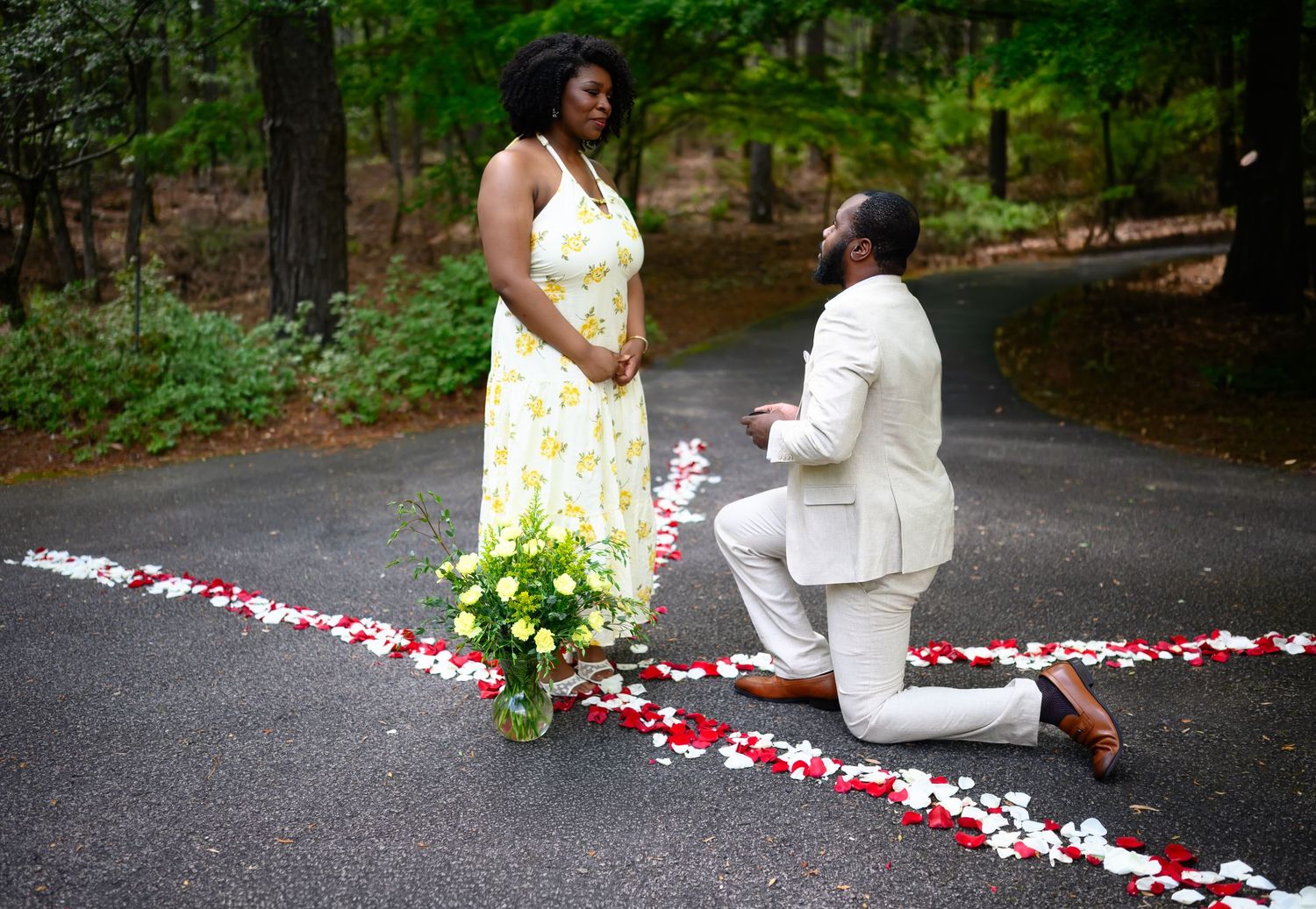 Man proposing on one knee to woman in yellow dress on a rose petal path in a wooded outdoor setting.