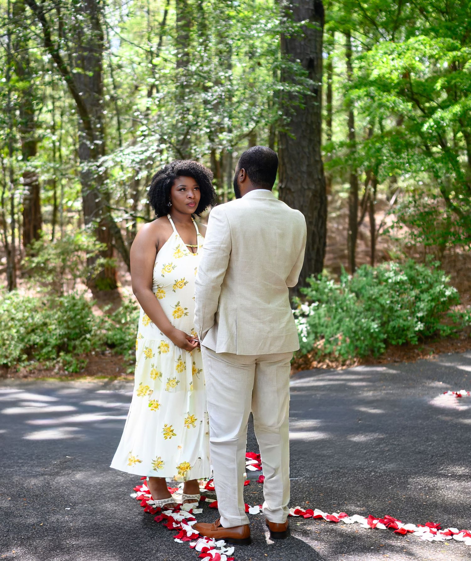 Couple stands back-to-back on a petal-lined path surrounded by tall green trees in a serene forest.