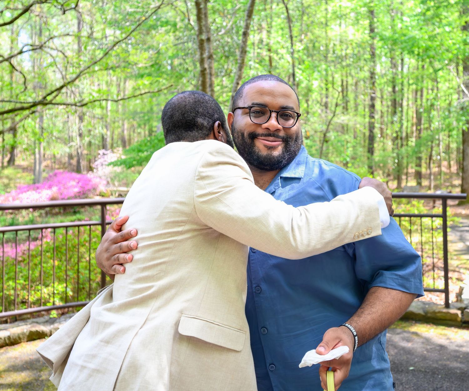 Two men share a warm congratulatory hug in a beautiful garden setting with pink azaleas blooming in the background.