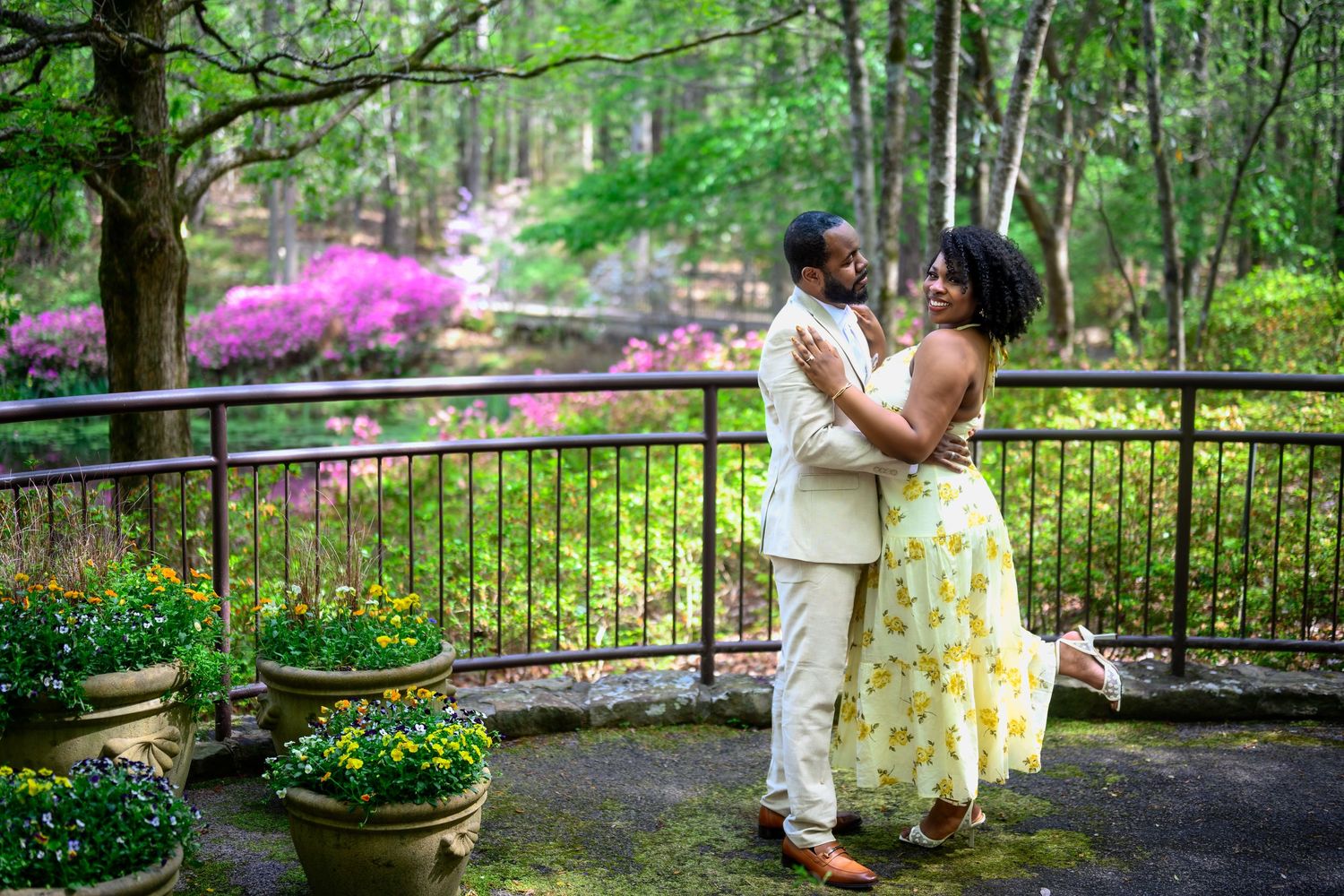 Joyful couple dancing and embracing on a garden terrace with vibrant pink azaleas and green trees behind them.