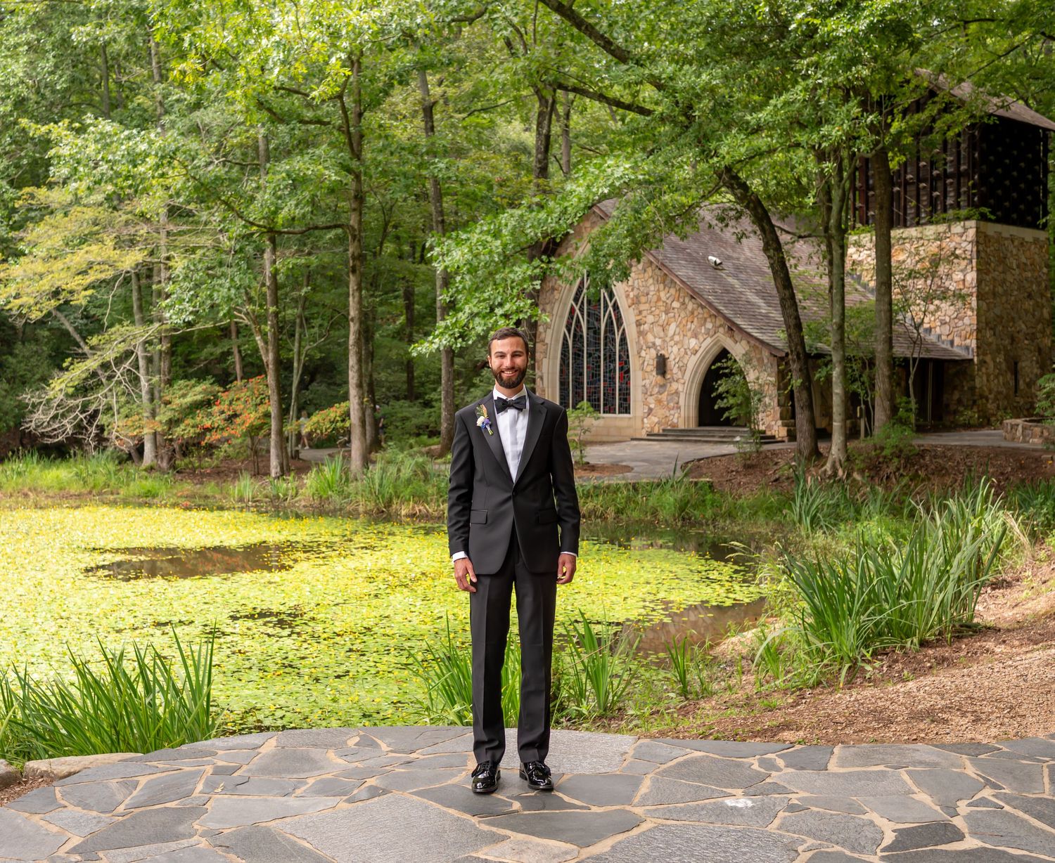A person in a black tuxedo stands on stone pavers in front of a rustic chapel nestled among lush green trees.