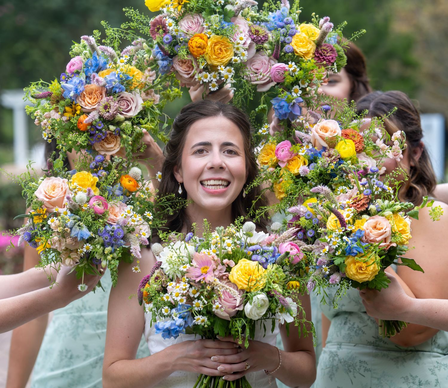 Three vibrant floral bouquets with yellow roses, pink blooms, and blue wildflowers are held up against a green outdoor background.