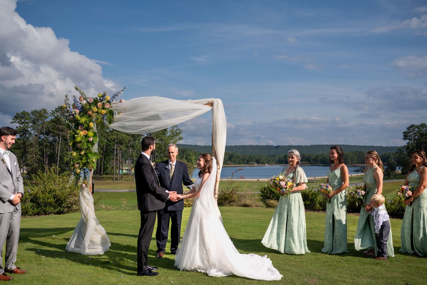 Outdoor wedding ceremony takes place under blue skies with guests gathered on a scenic grassy lawn by the water.