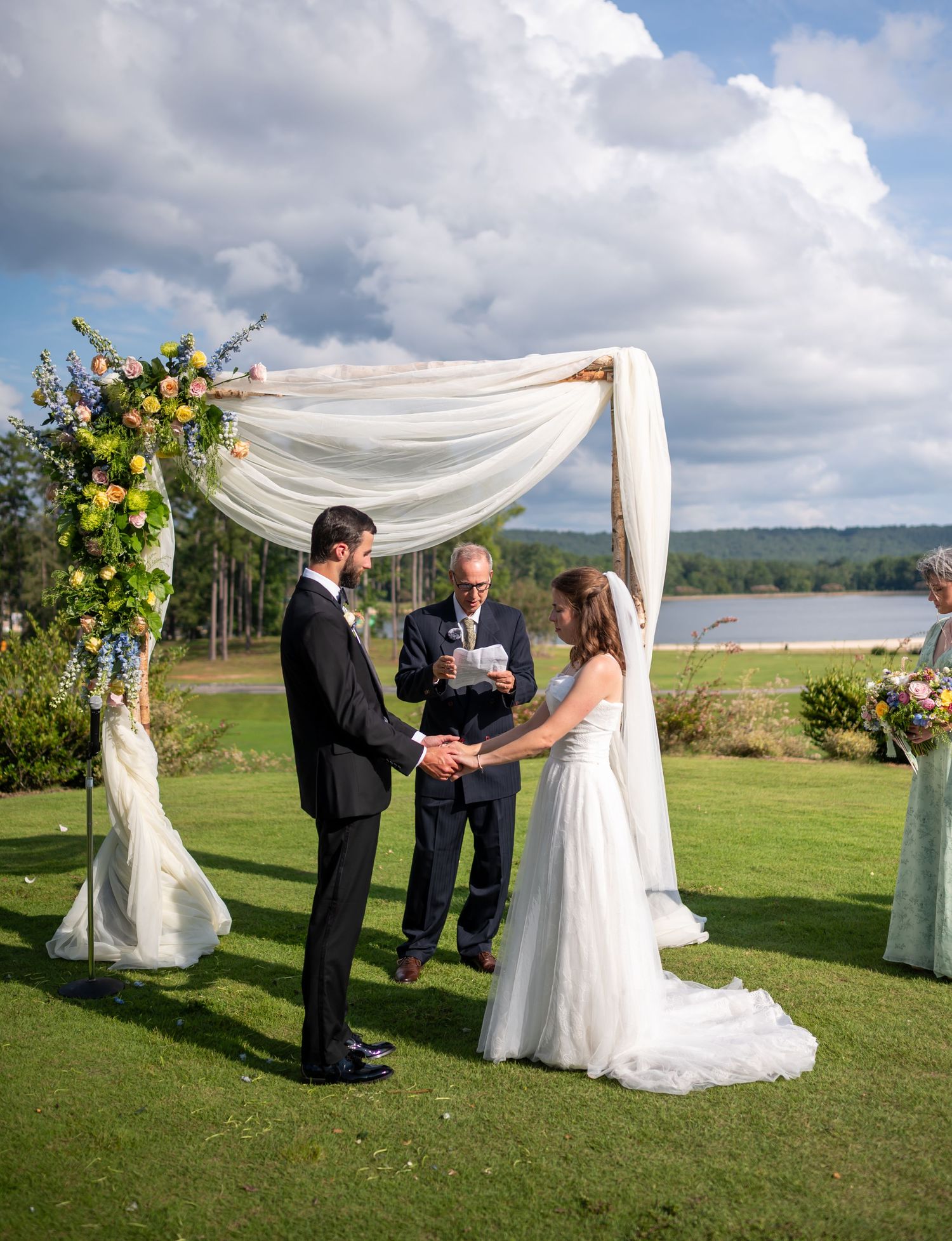 A wedding ceremony takes place on a scenic waterfront lawn with white fabric draped archway and floral arrangements.