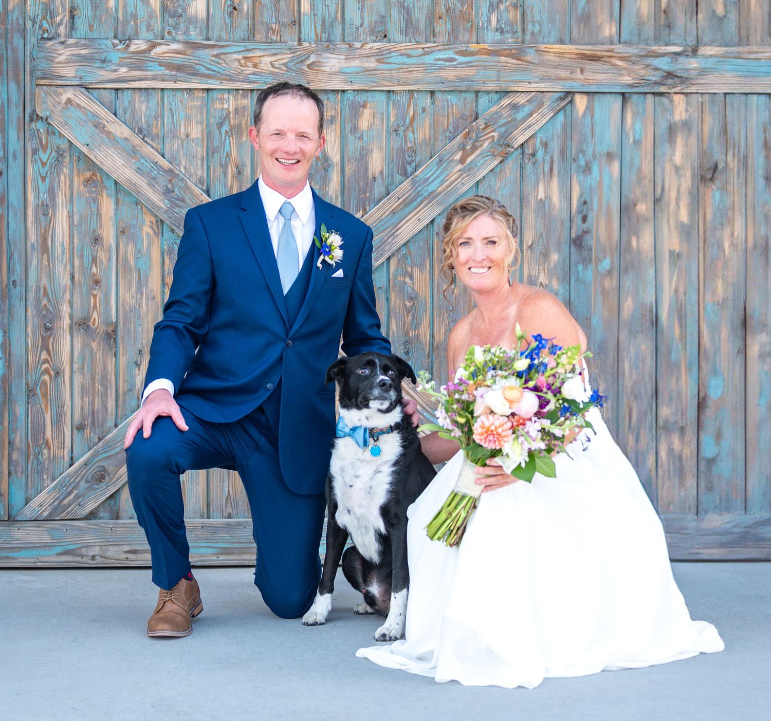 A wedding portrait featuring a couple and their black and white dog in front of a weathered blue barn door.
