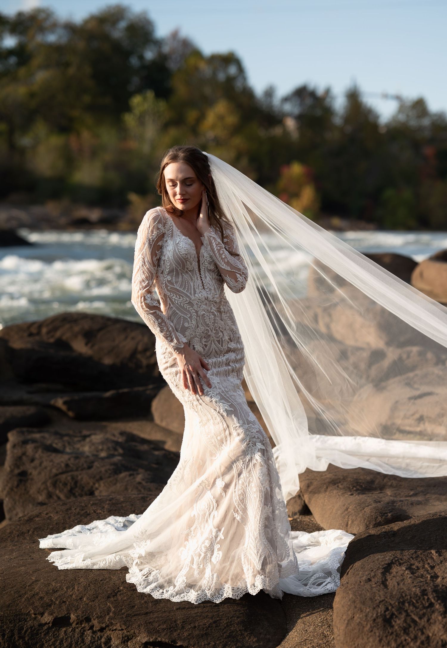 Fitted lace wedding gown with long sleeves and cathedral veil photographed on rocky riverbank.
