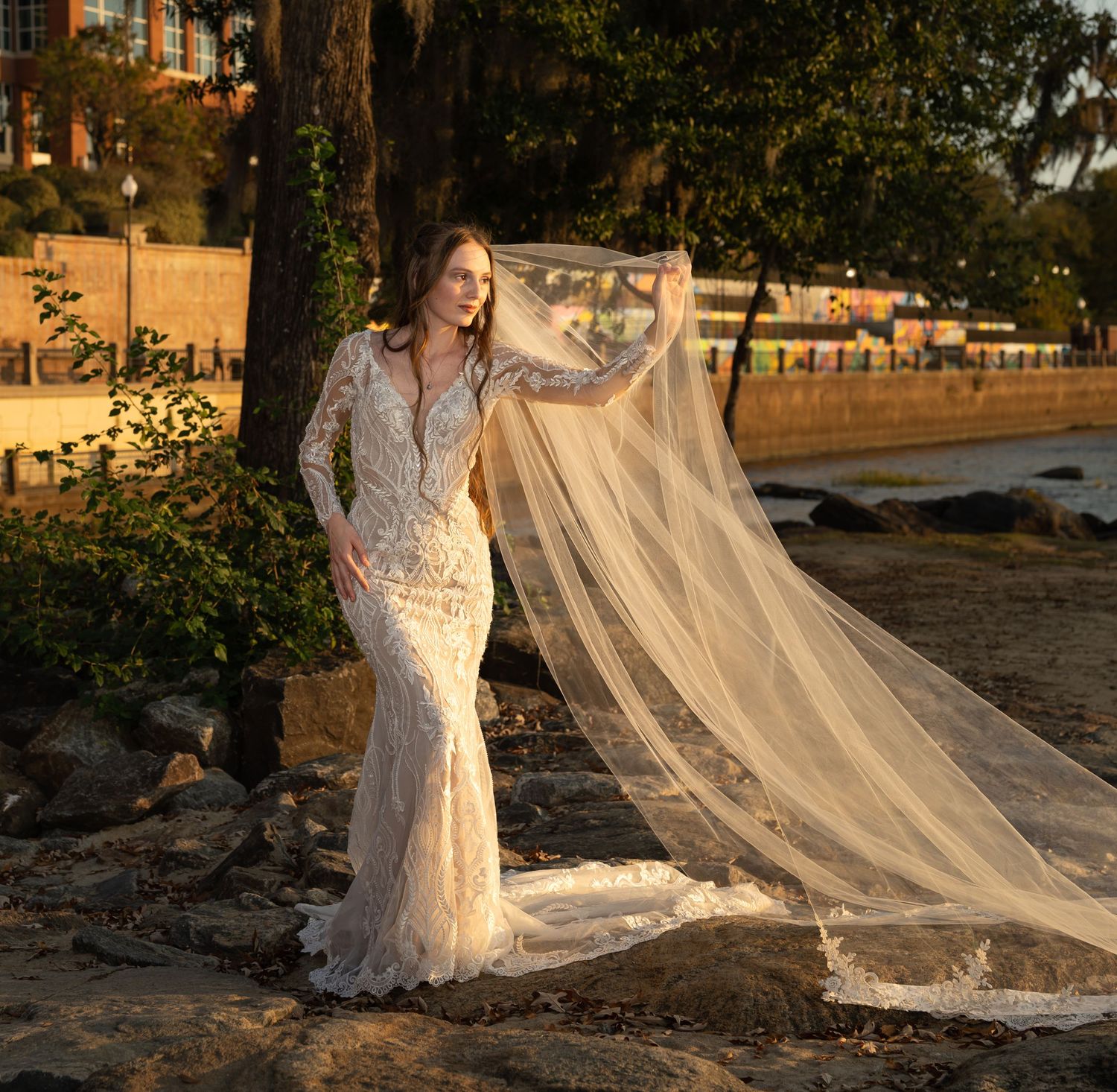 A bridal portrait at sunset showing a fitted lace gown with dramatic veil against brick architecture.