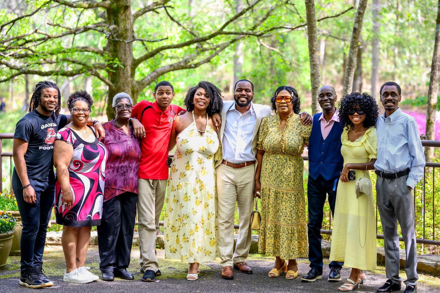 A large joyful family group poses together outdoors in a lush green park celebrating an engagement announcement.