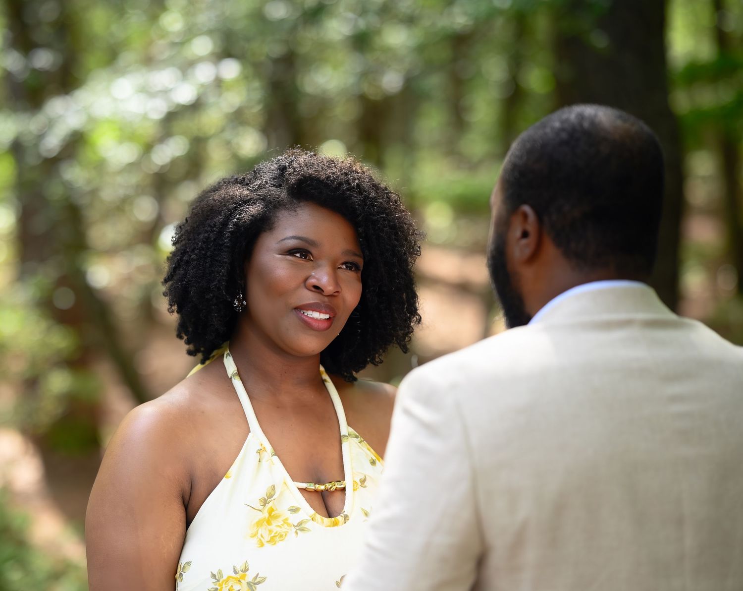 A woman in a yellow floral halter dress smiles at a man in a beige suit in a wooded setting.