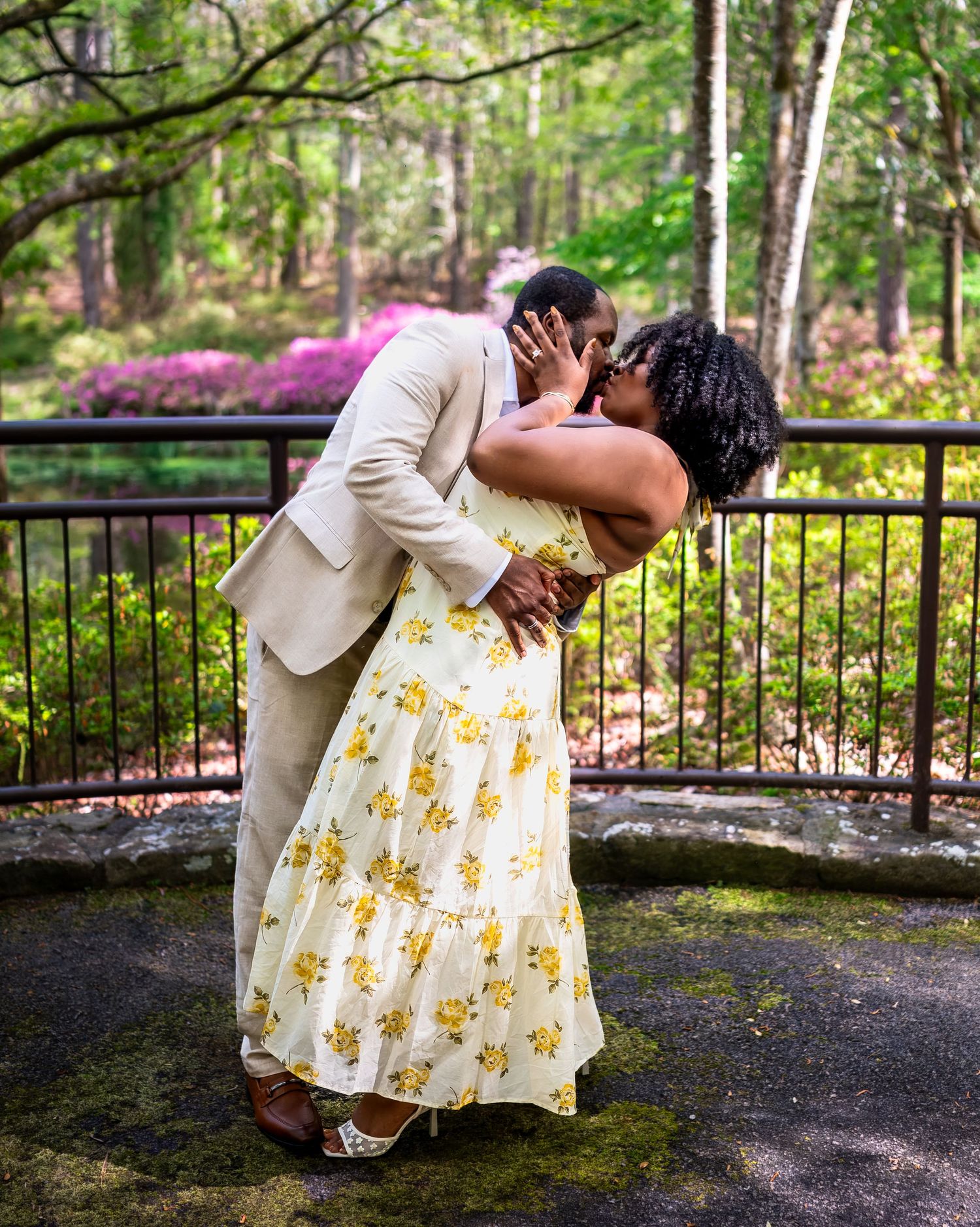 A couple sharing a passionate dip kiss outdoors surrounded by lush greenery and blooming azaleas.