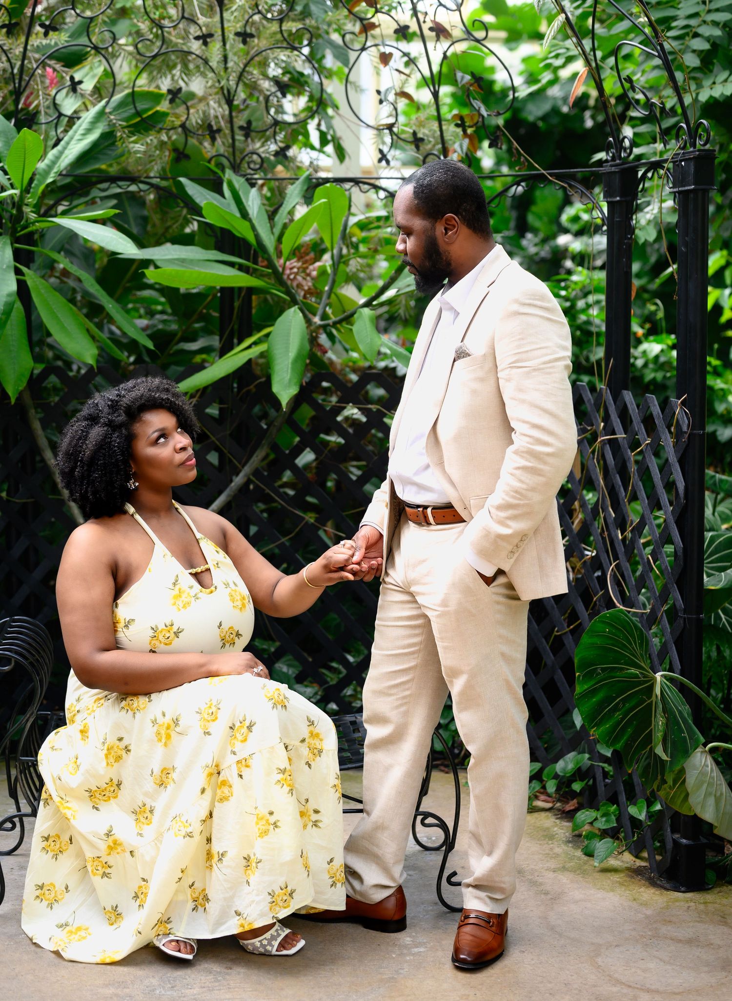 Couple holding hands and gazing at each other among tropical plants and iron gates.