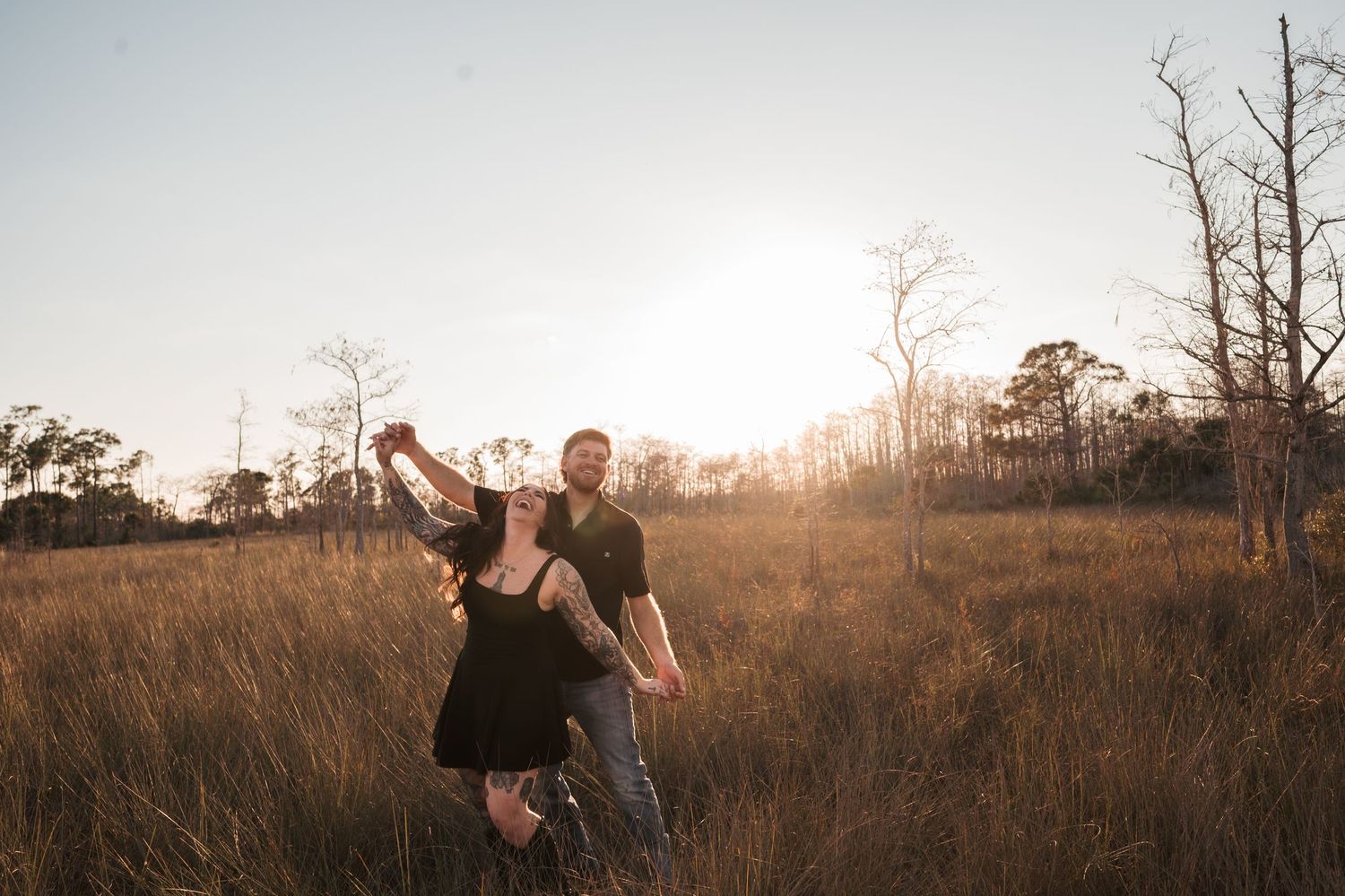 A couple embraces in a golden field during sunset while surrounded by tall grass and bare trees.