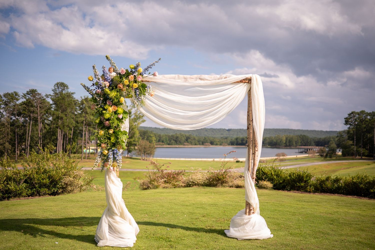 A scenic waterfront wedding ceremony setup with white fabric draped arch and chairs overlooking a serene lake.