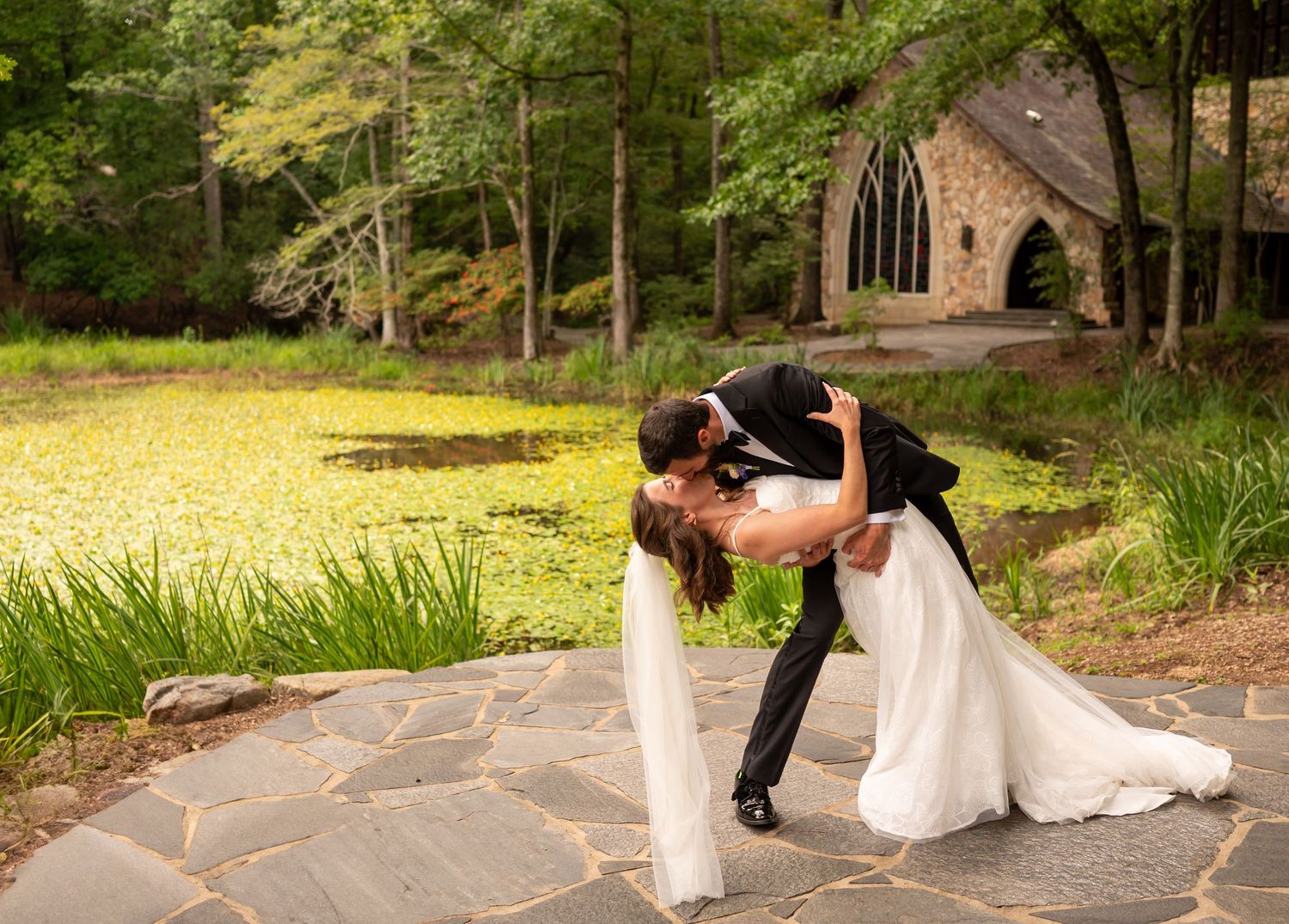 A romantic wedding couple dips in a choreographed dance pose on a stone path surrounded by lush green forest.
