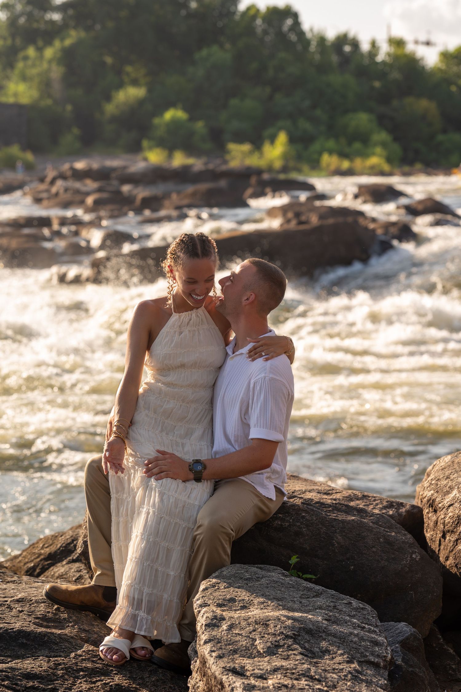 Couple sits on rocky riverbank with rushing white water rapids in background during sunset.
