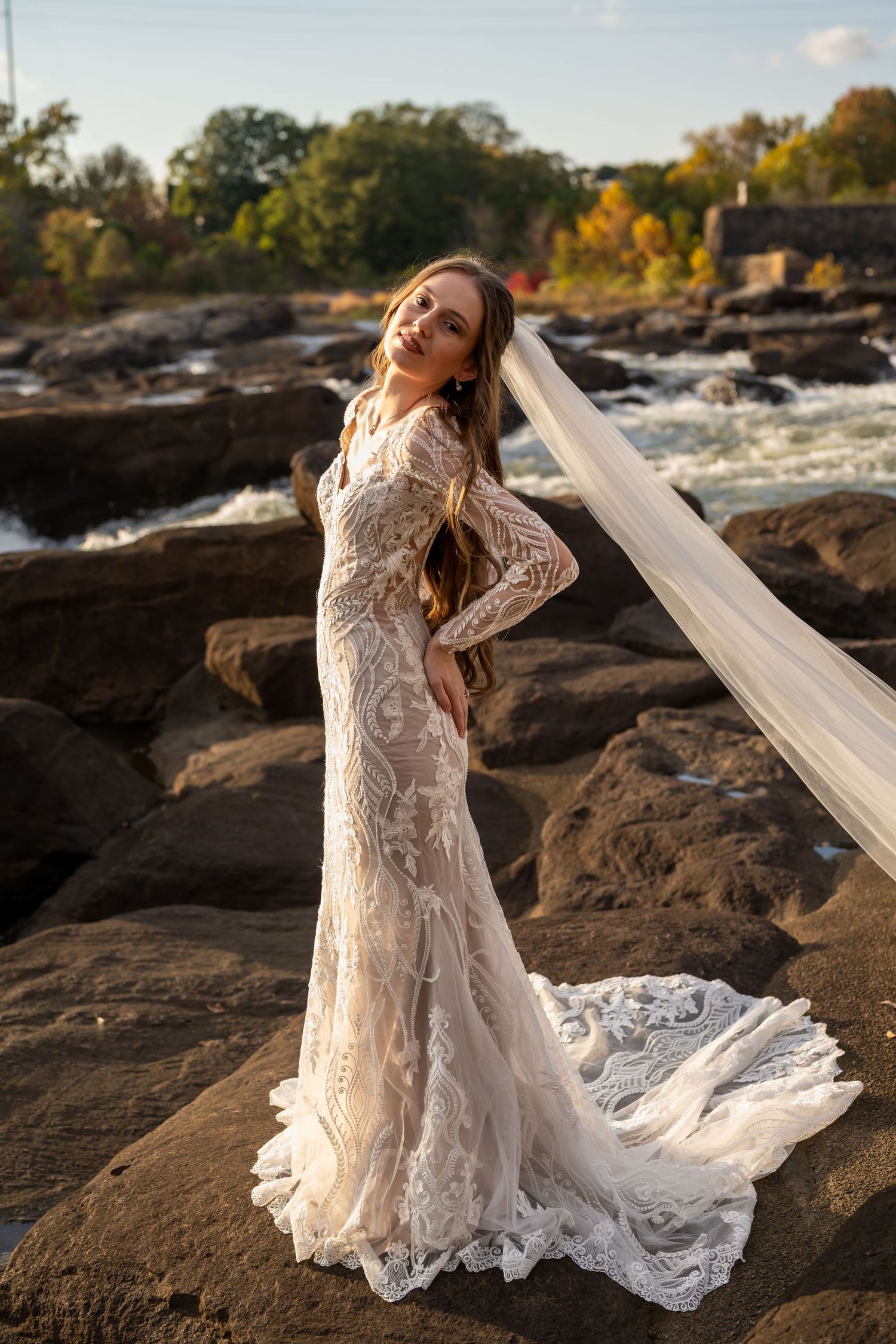 A flowing white wedding dress with lace details cascades over dark rocks near rushing water.