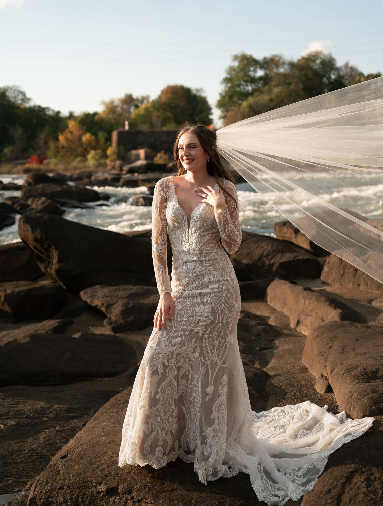 Long sleeve lace wedding dress with flowing veil photographed on rocky coastline during golden hour.