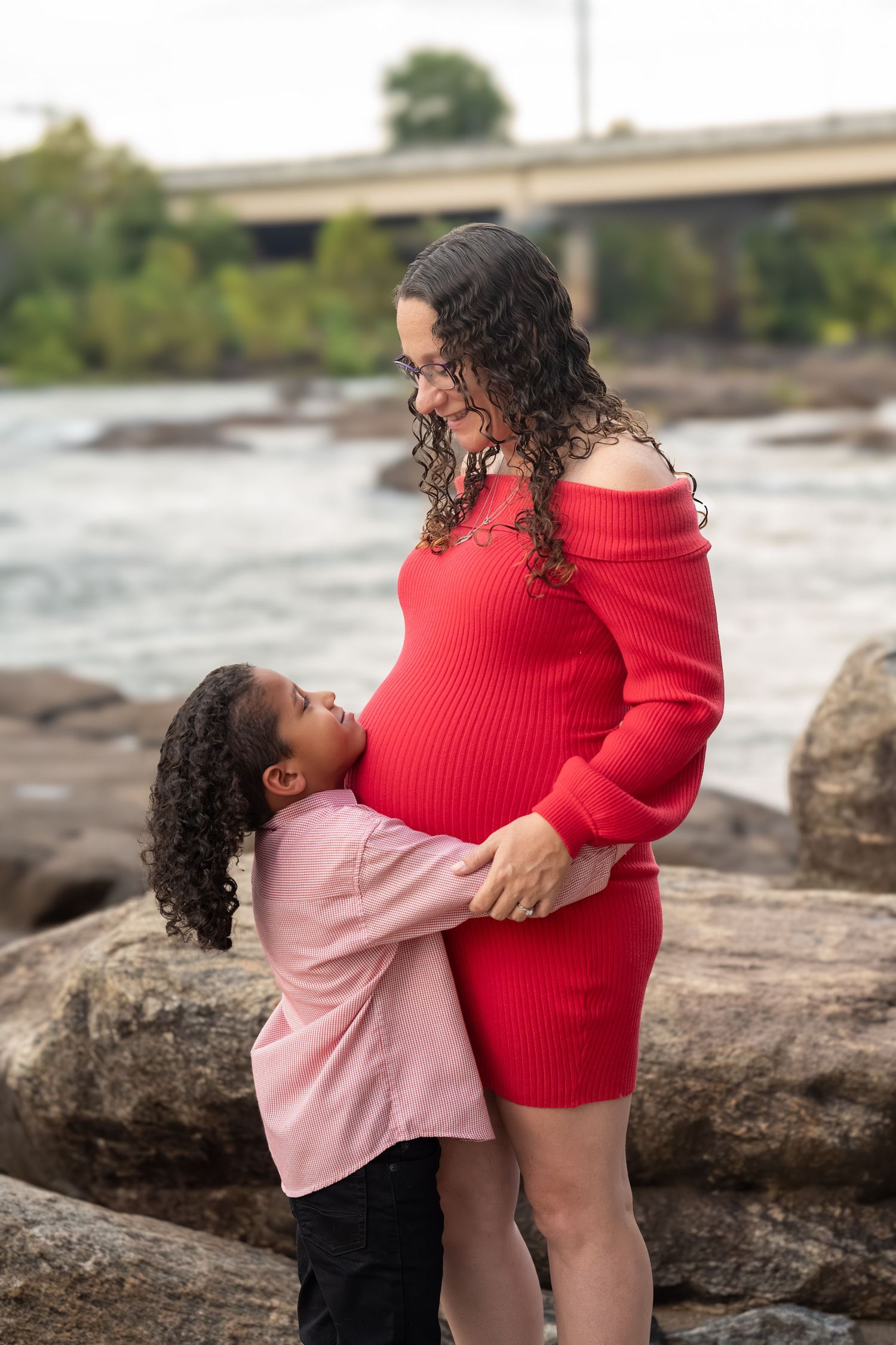 A tender maternity moment captured on rocky terrain as a young child embraces an expectant mother in a red dress.