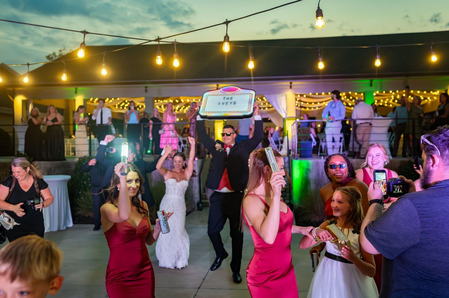 Wedding guests dance and celebrate on an outdoor patio decorated with string lights and colorful decor at dusk.