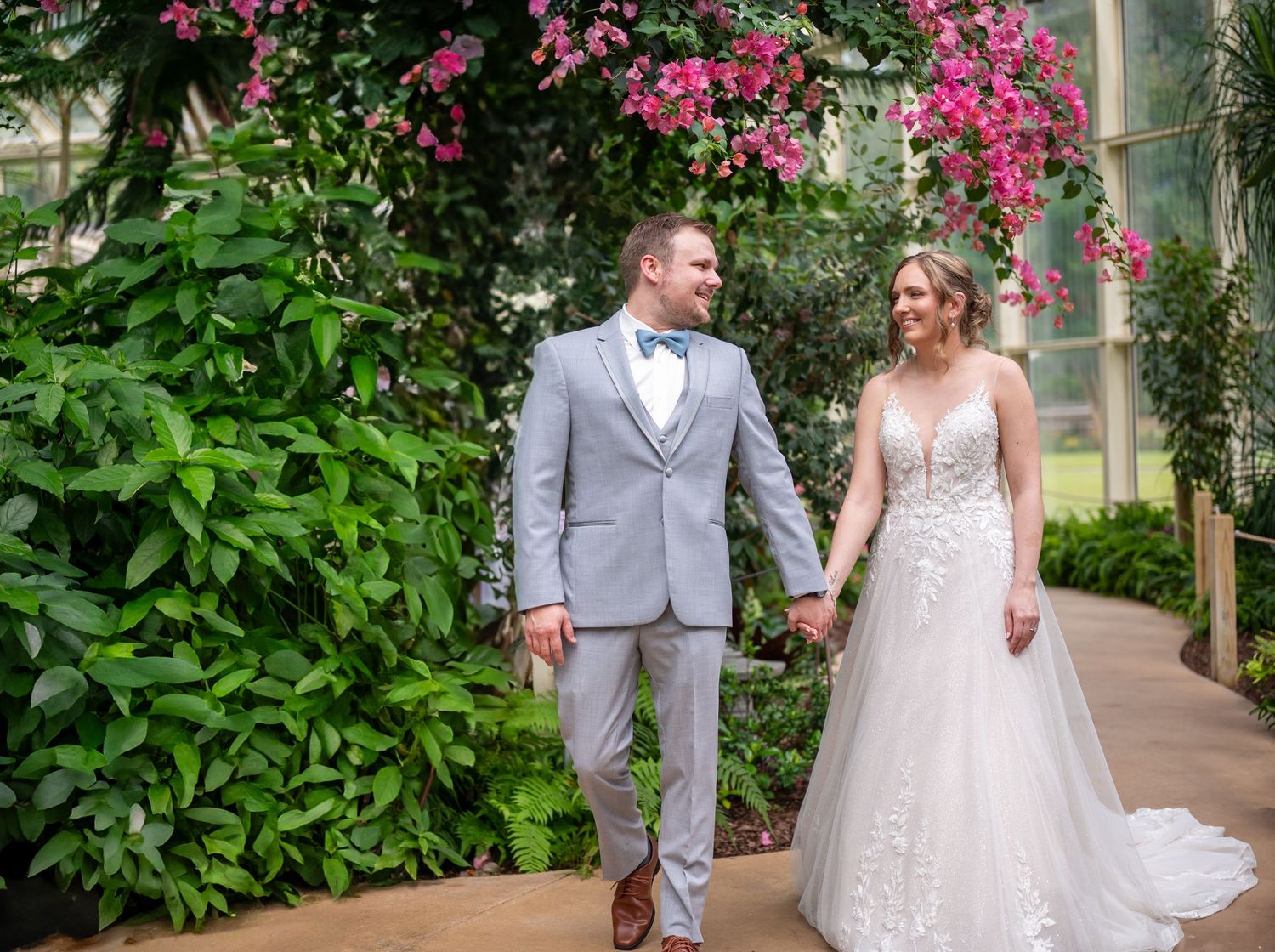 Wedding couple walking along garden path surrounded by vibrant pink flowers and tropical greenery.