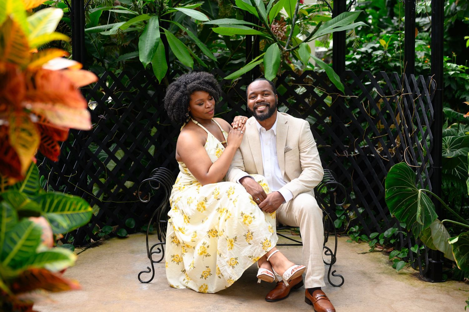 Smiling couple poses playfully on a garden bench surrounded by rich green tropical foliage.