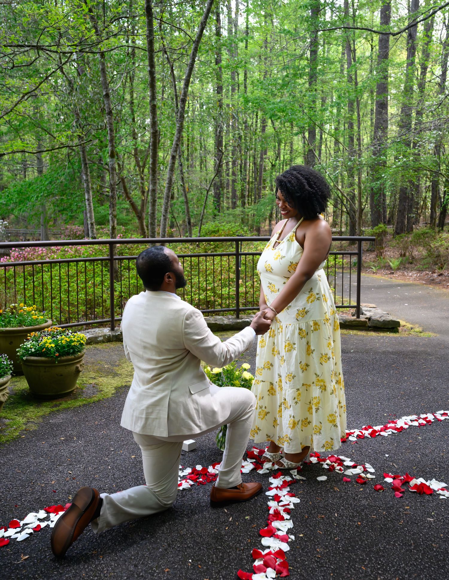 Man proposes on one knee to woman in yellow floral dress surrounded by rose petals in a wooded outdoor setting.