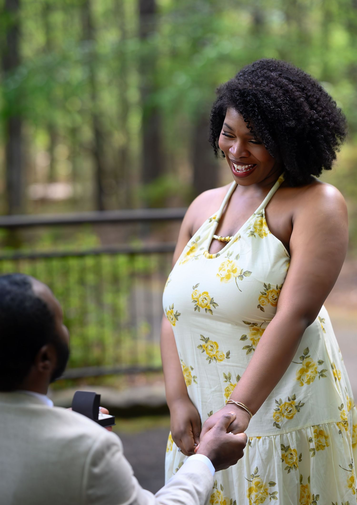 Joyful woman in yellow floral dress smiling as man kneels proposing on woodland path with rose petals.