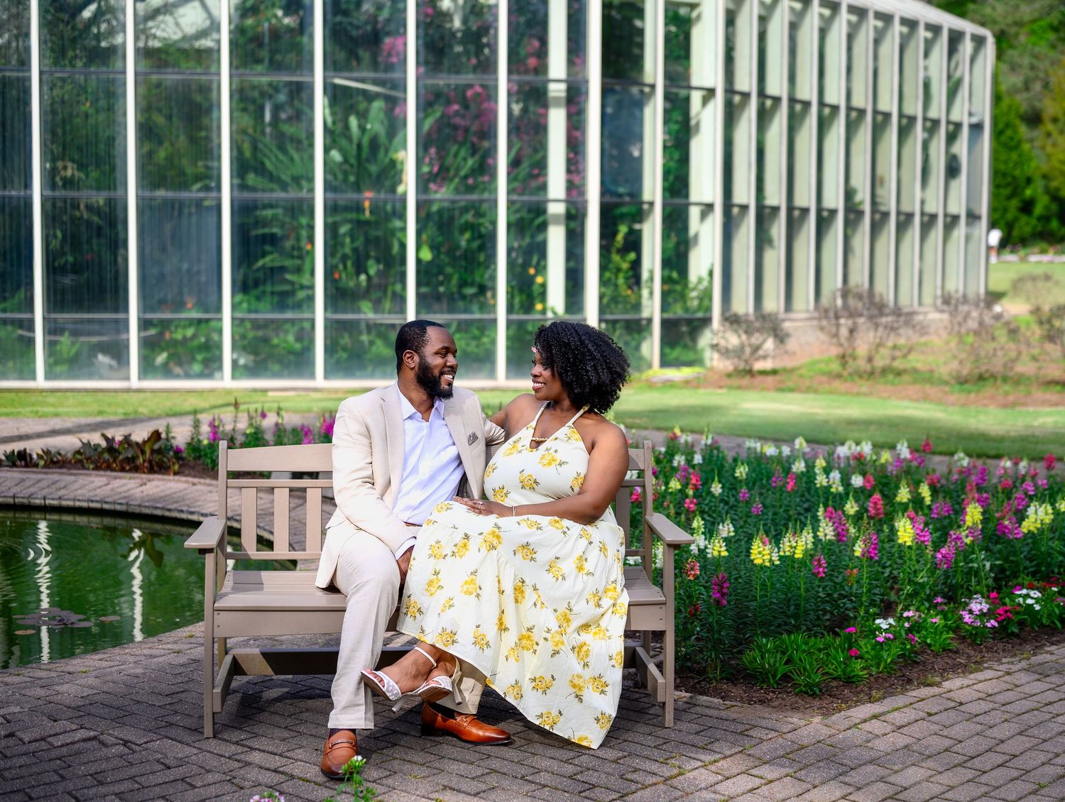 A couple sitting on a wooden bench by a pond, surrounded by colorful tulips near a greenhouse.