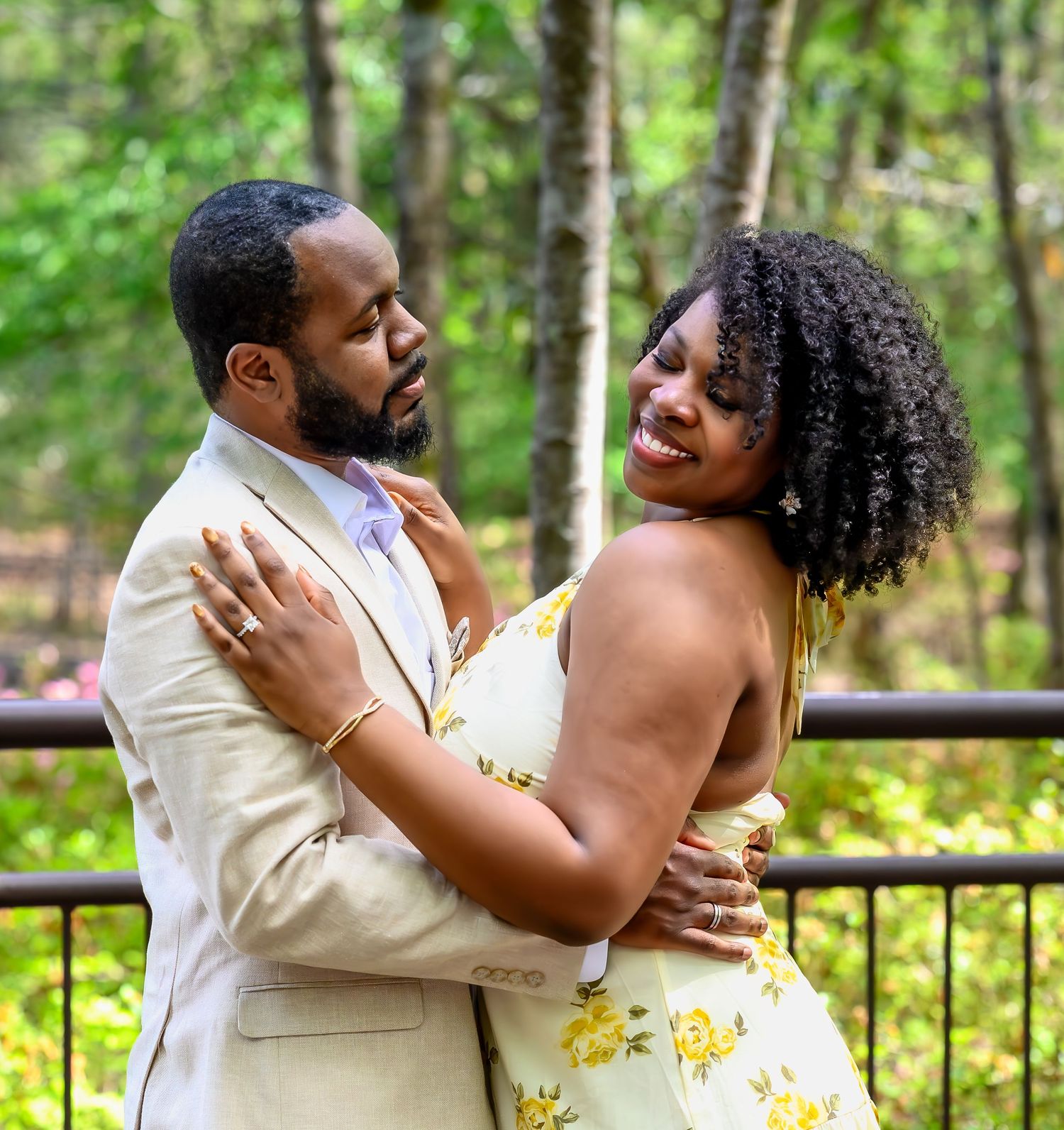 A happy engaged couple embraces on a wooden bridge surrounded by lush green trees in a park setting.