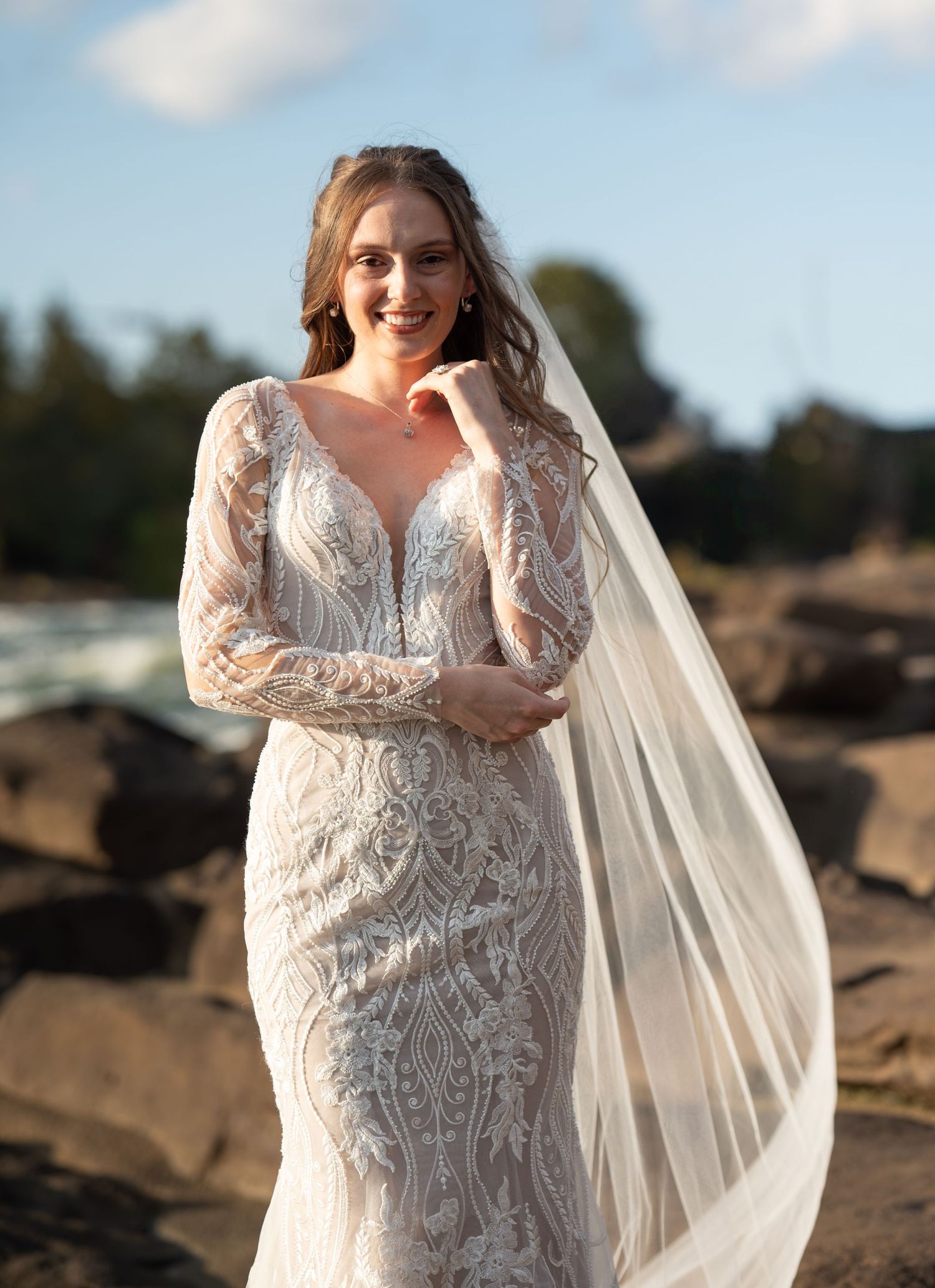 A bride in a lace wedding gown poses gracefully on rocky terrain during a sunset photoshoot.