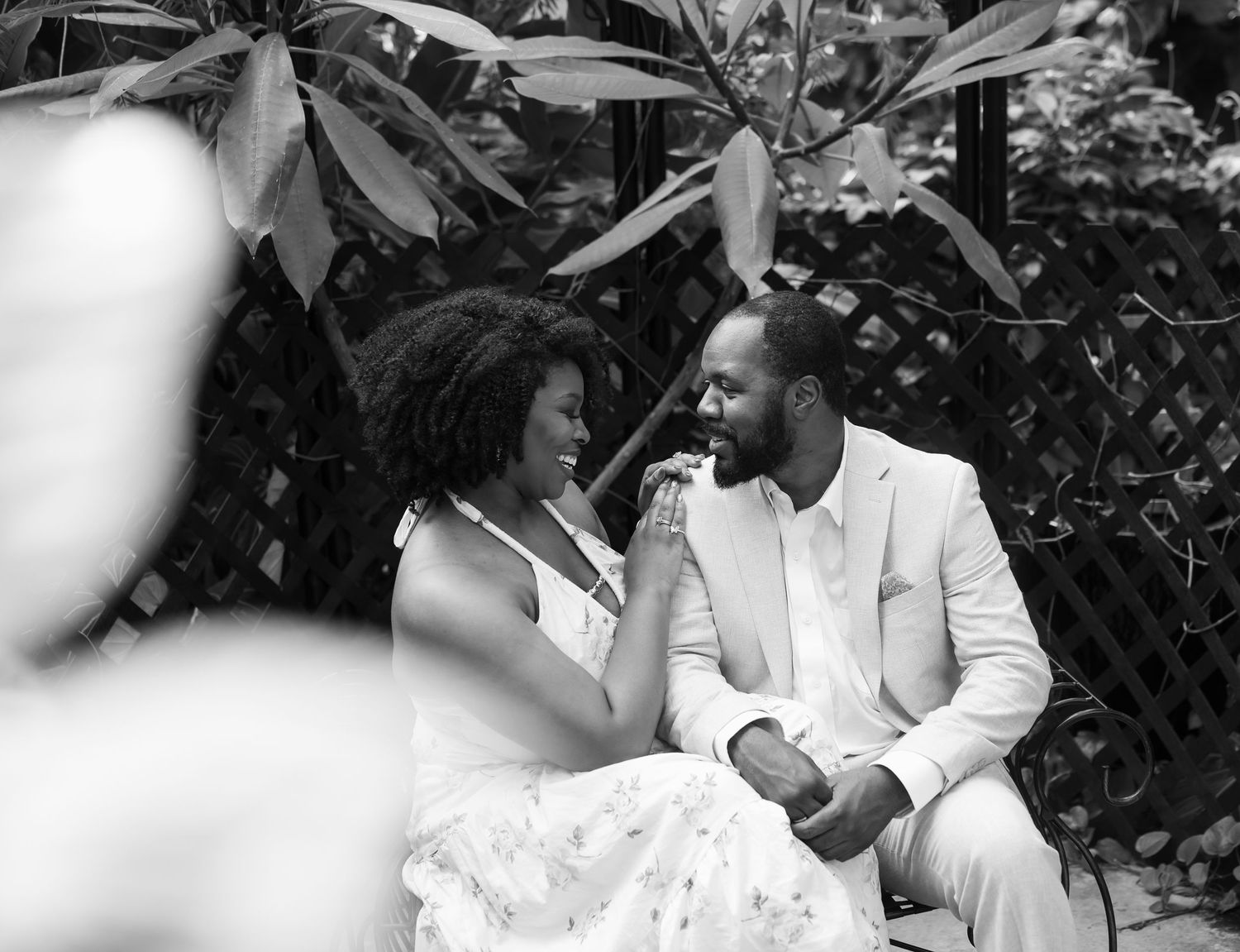Black and white photo of a couple sitting closely together smiling among tropical garden foliage.