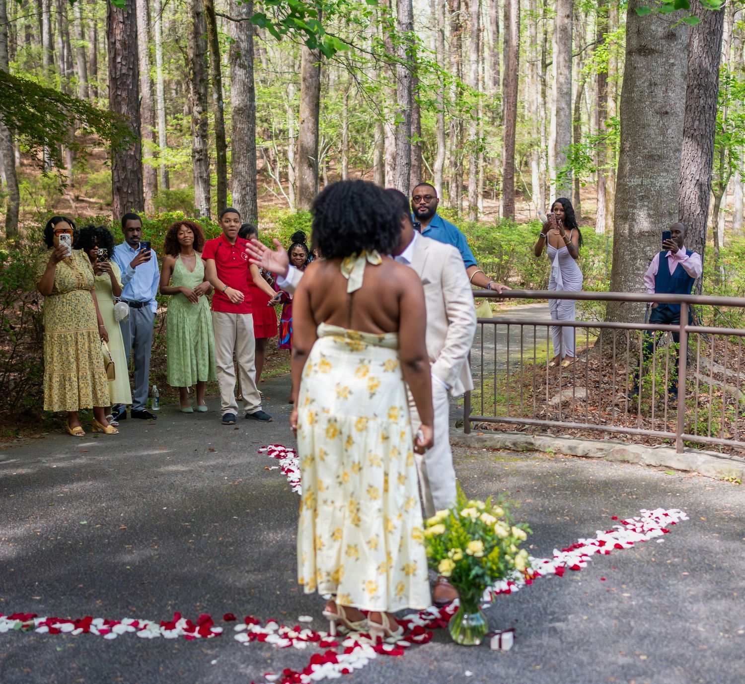 Outdoor woodland marriage proposal with guests watching as couple stands on rose petal path in forest setting.