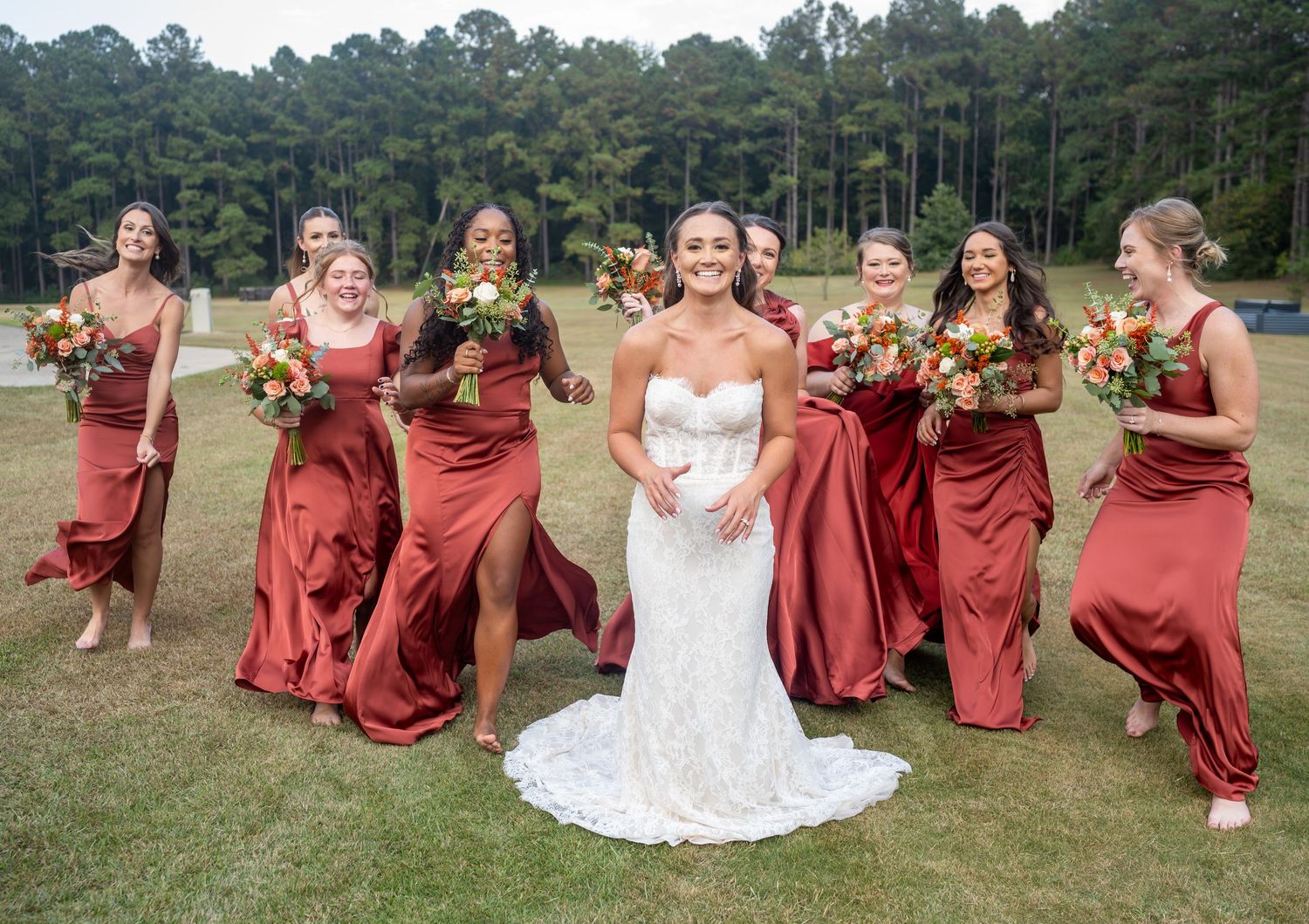 Wedding party in rust-colored dresses and white gown pose joyfully on grass with pine trees in background.