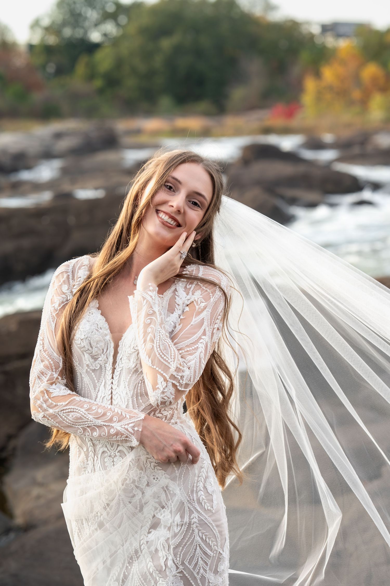 A flowing white lace wedding dress and veil catch the sunlight on rocky terrain near water.