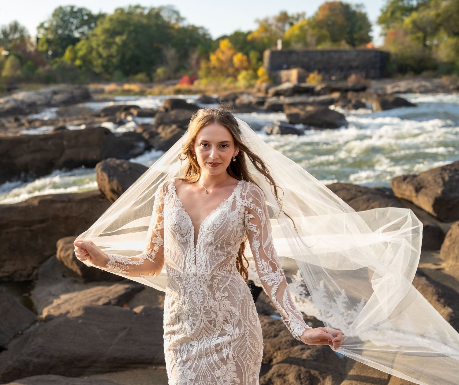 Wedding dress with intricate lace details and flowing veil captured against autumn riverside scenery.