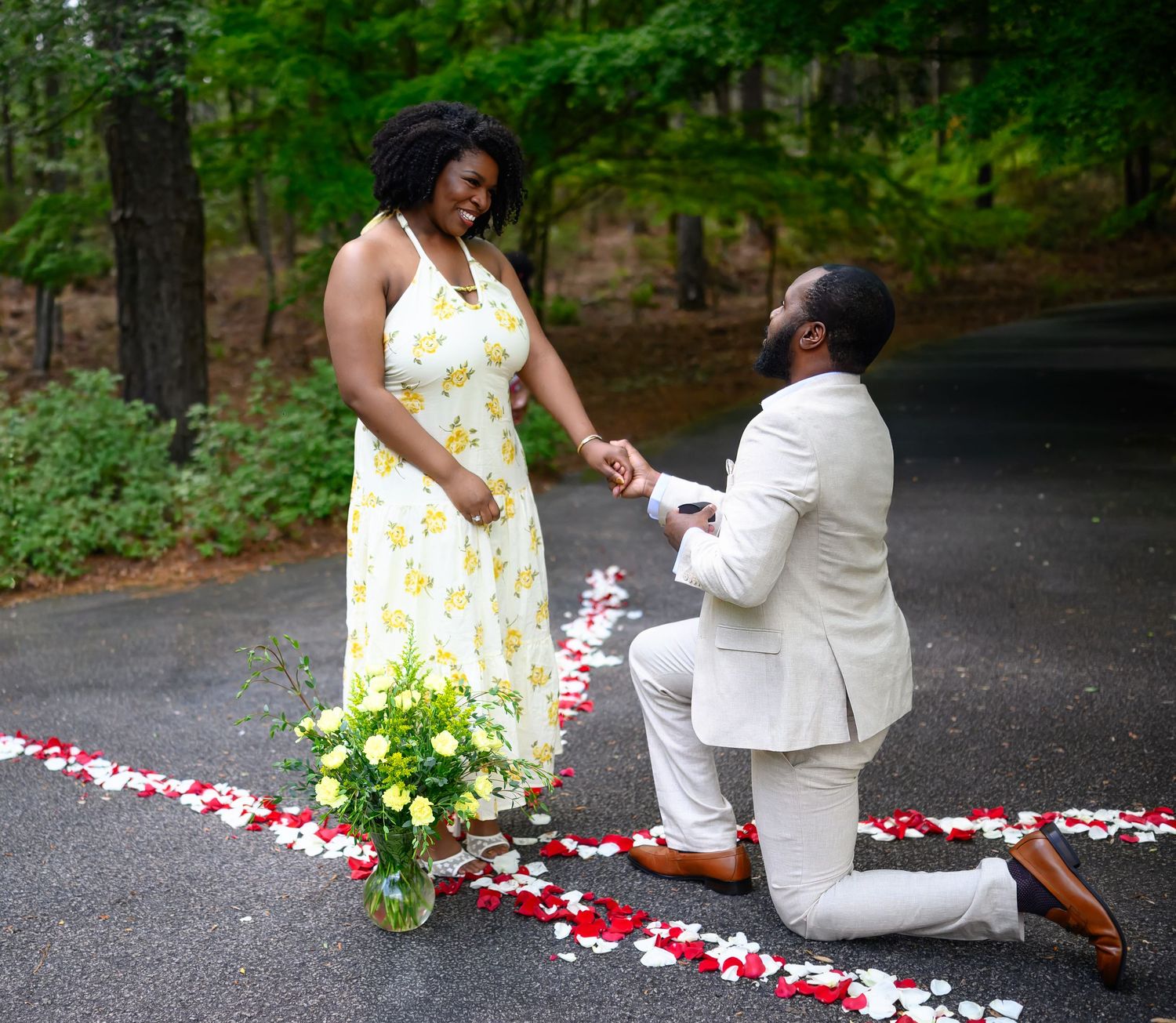 Man in cream suit proposes on one knee to smiling woman in yellow floral dress amid rose petals.