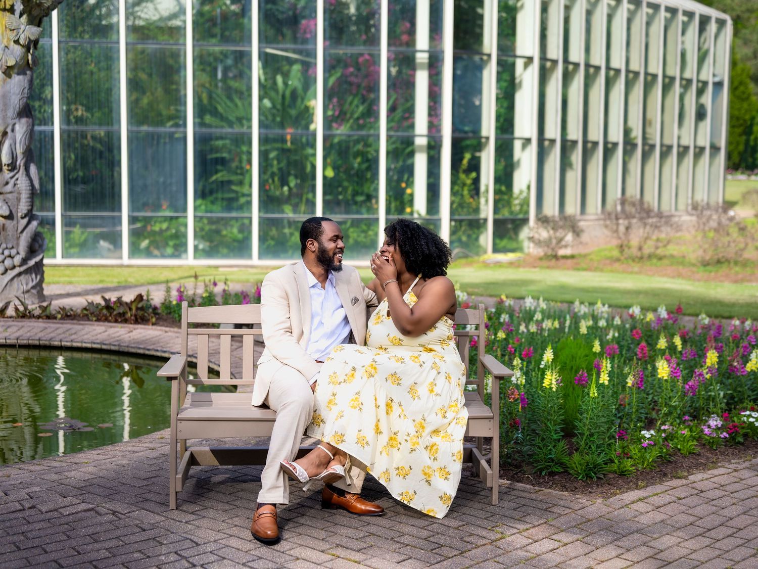 A couple shares a joyful moment seated on a bench near a garden pond with colorful flowers.