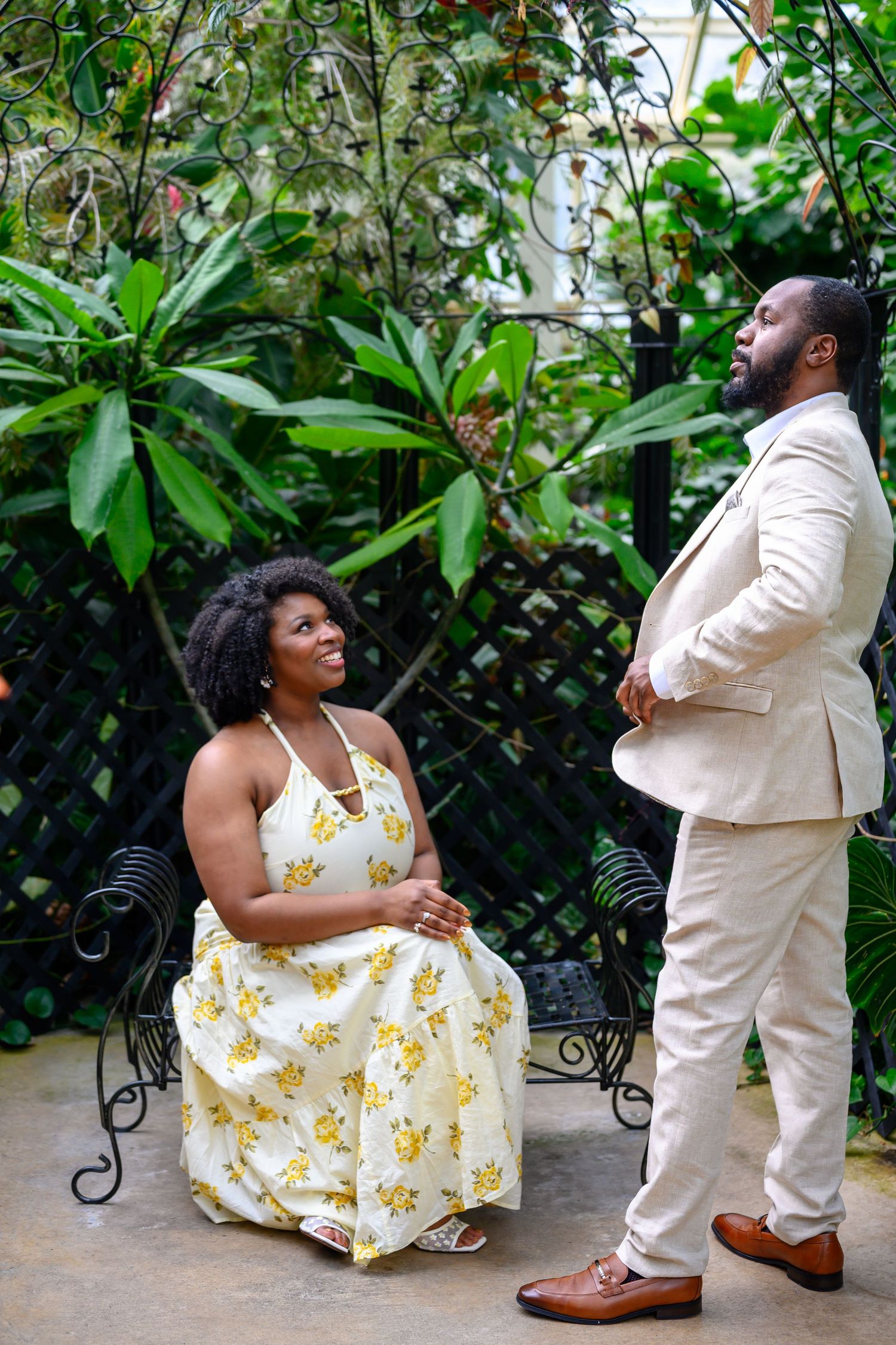 A woman in a yellow floral dress sits on an iron bench smiling up at her partner in a beige suit.