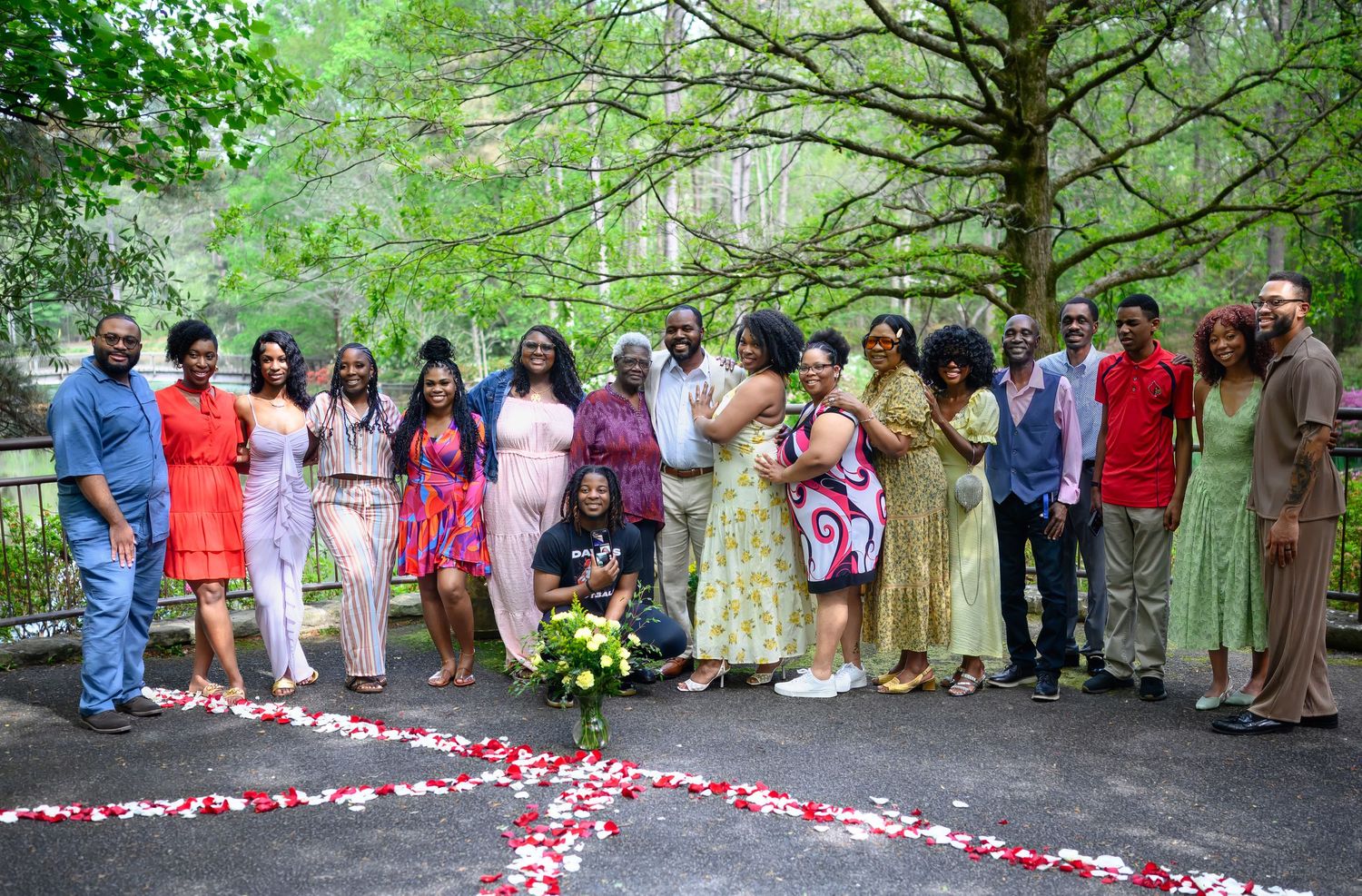 Large family group celebrates engagement surrounded by rose petals on outdoor patio in wooded area.