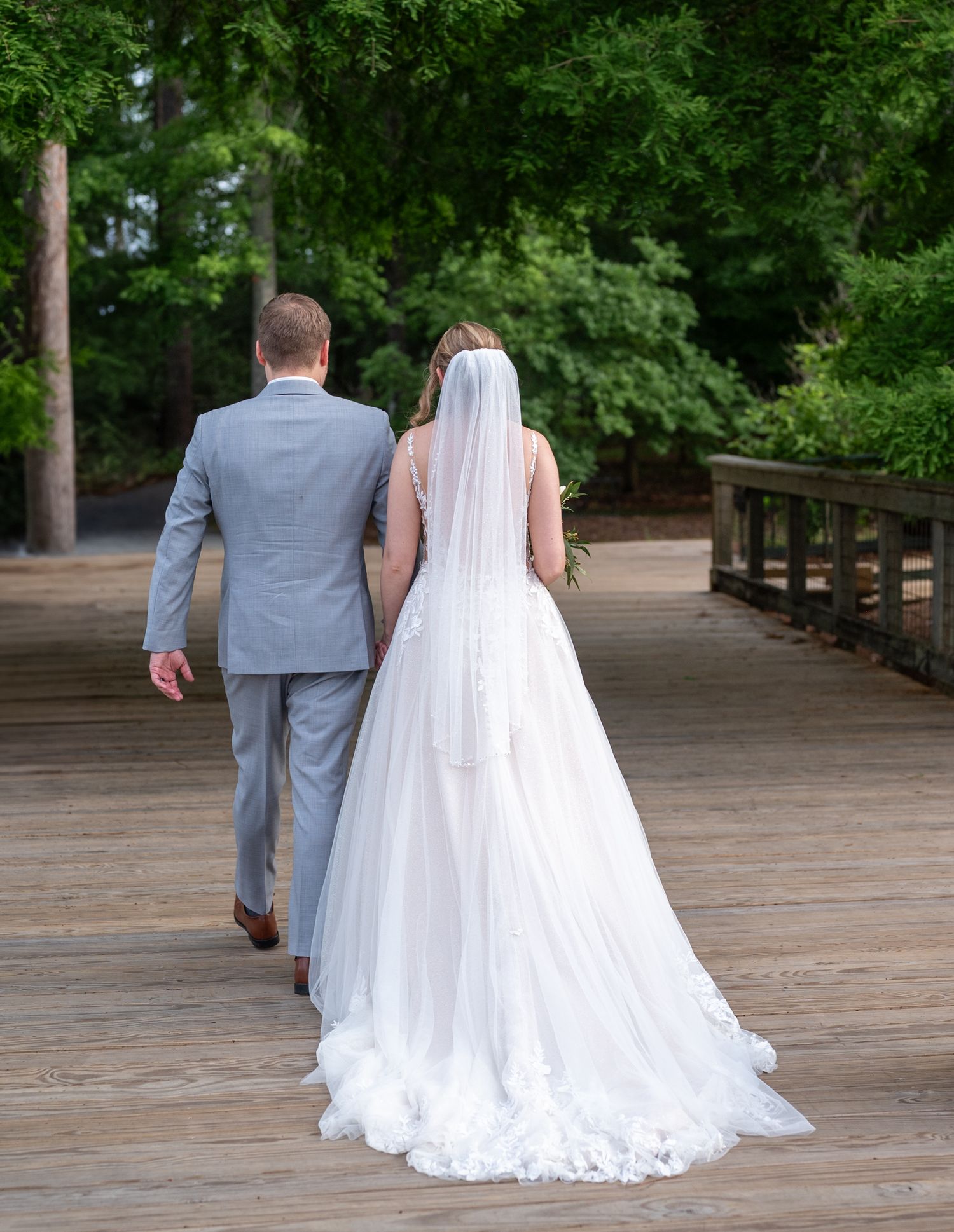 A couple in wedding attire walk down a scenic wooden boardwalk surrounded by lush greenery.