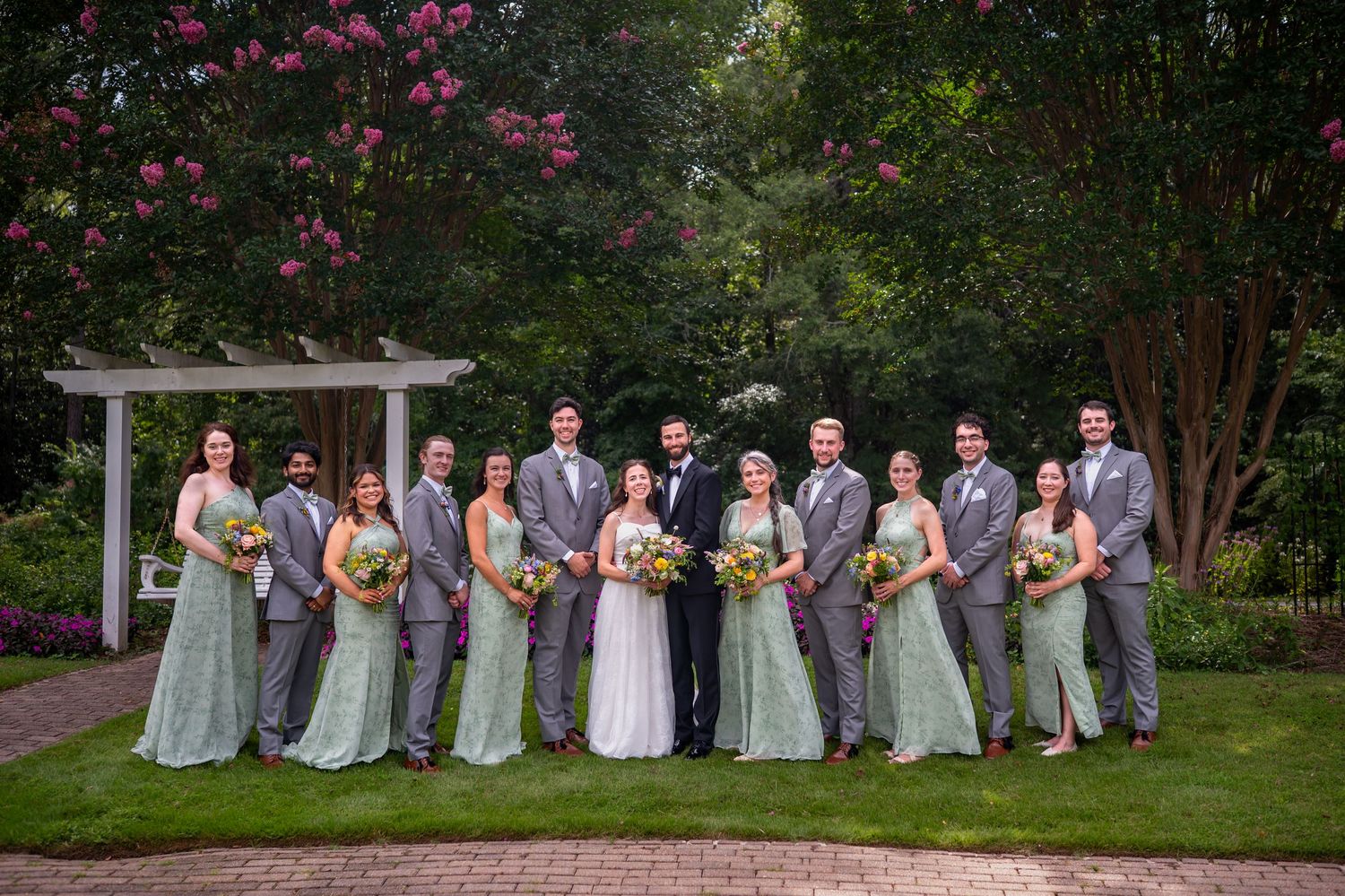 Wedding party in light gray dresses poses on lush garden lawn near white gazebo.