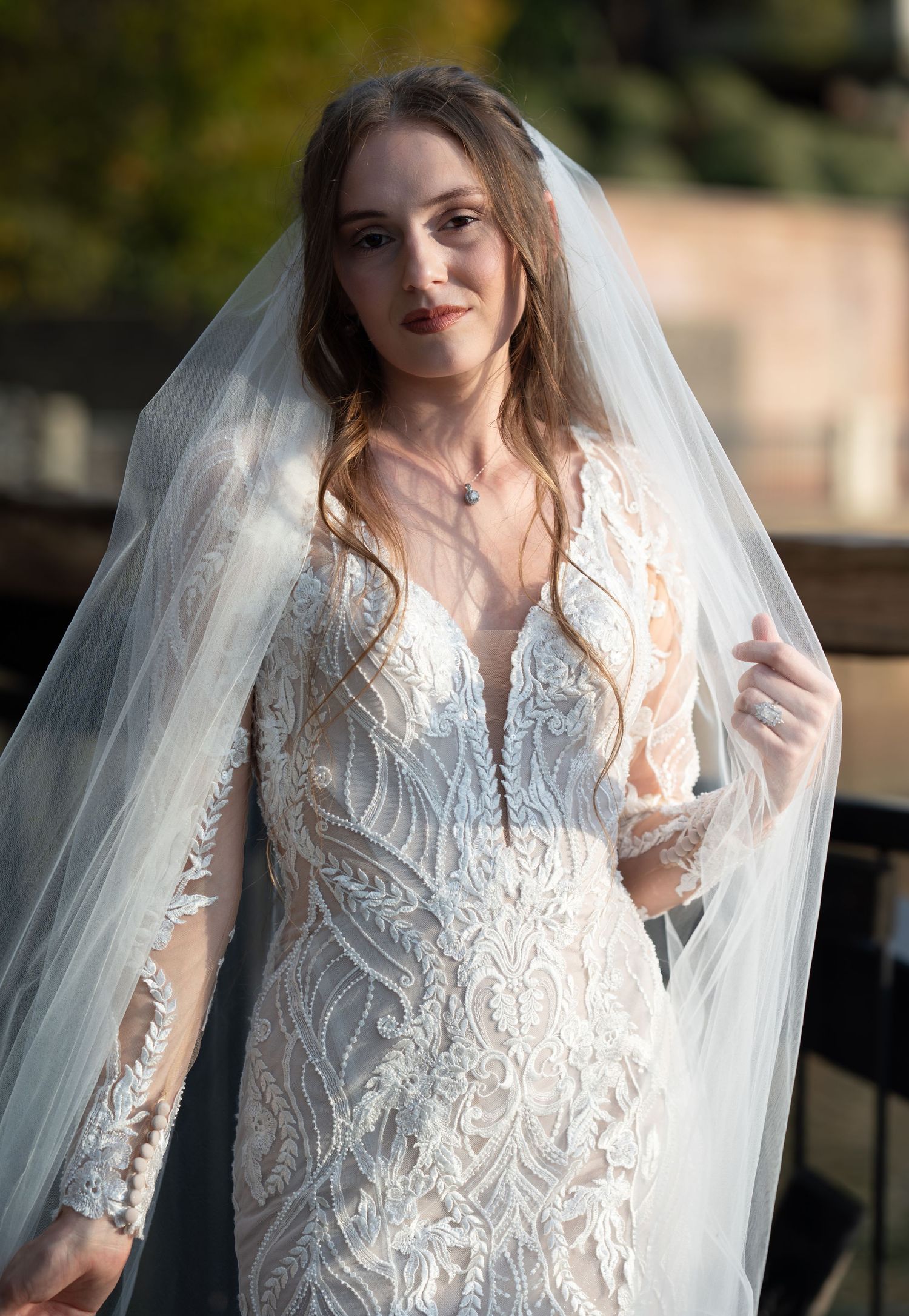Delicate lace wedding gown with sheer sleeves and plunging neckline shown in romantic outdoor portrait.
