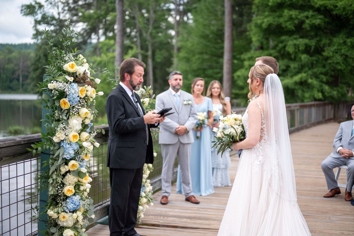 Wedding ceremony in progress on wooden dock with floral arch and lake backdrop.