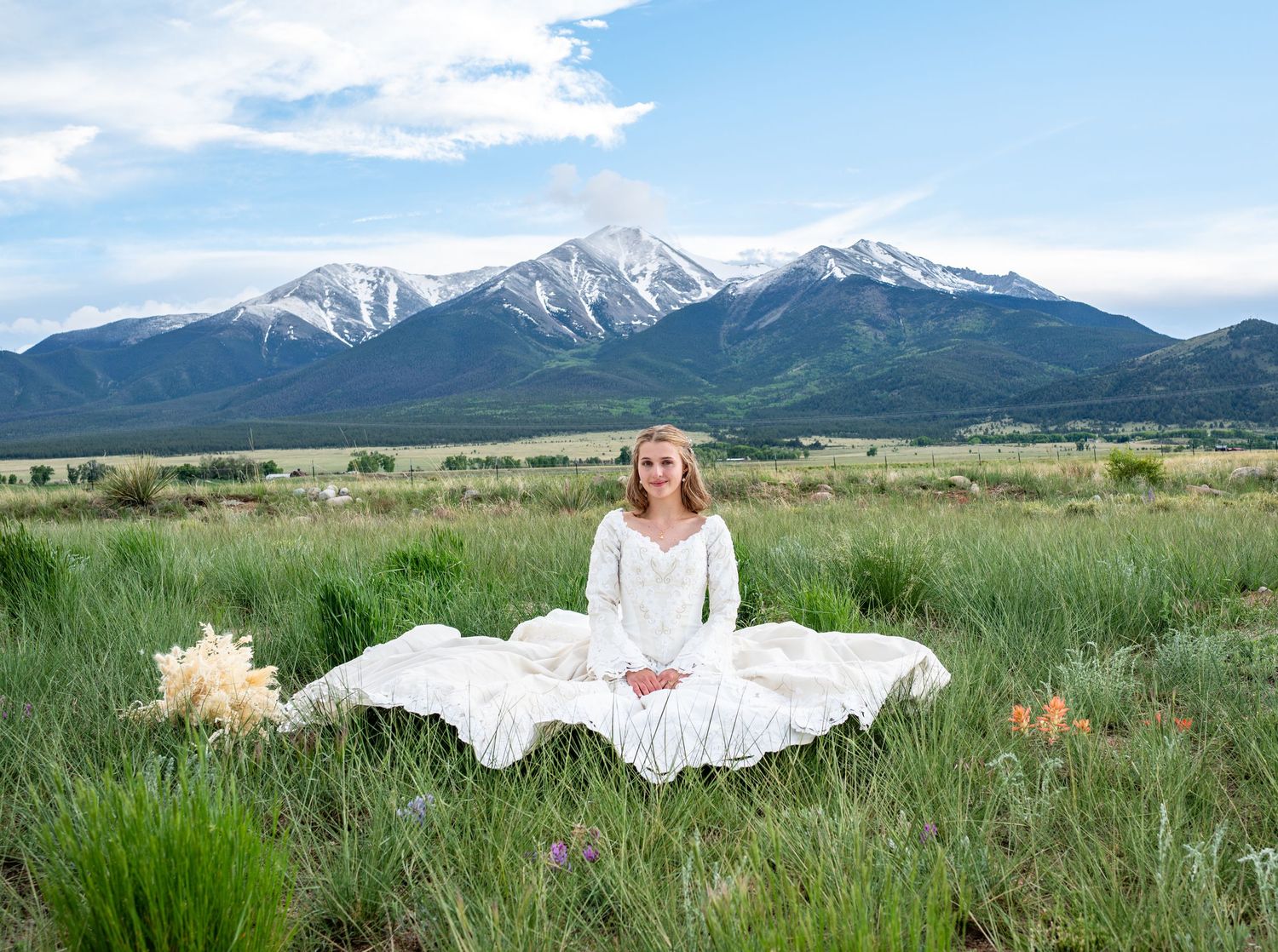 Bride in a vintage wedding gown sitting in the grass with wildflowers present. Huge 14,000 ft mountain behind her in Buena Vista, Colorado