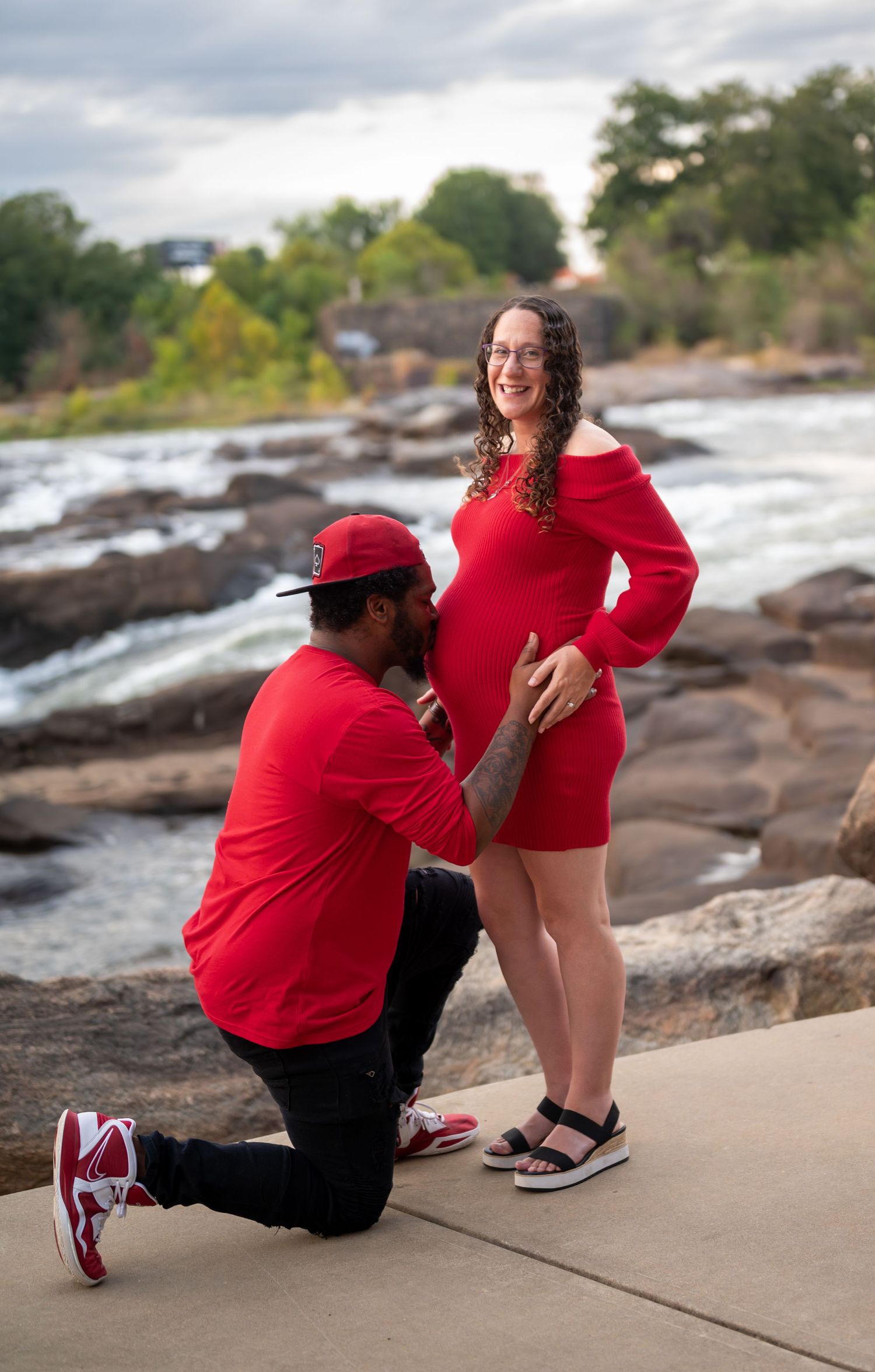 A couple in matching red outfits poses on rocky terrain near a waterfront during an outdoor photoshoot.