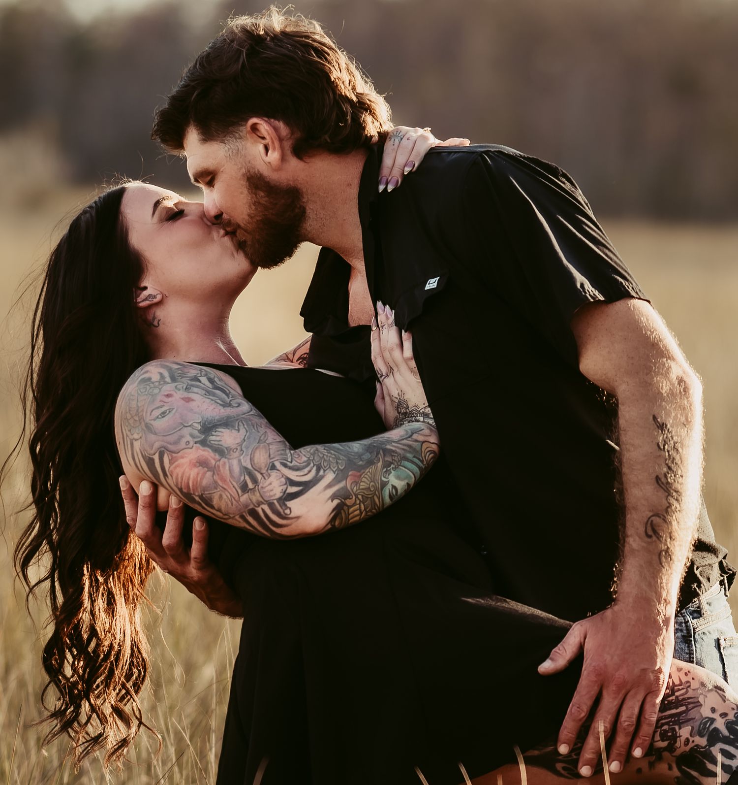 Romantic couple sharing an intimate kiss in a sunlit field, with one person displaying artistic tattoo sleeves.