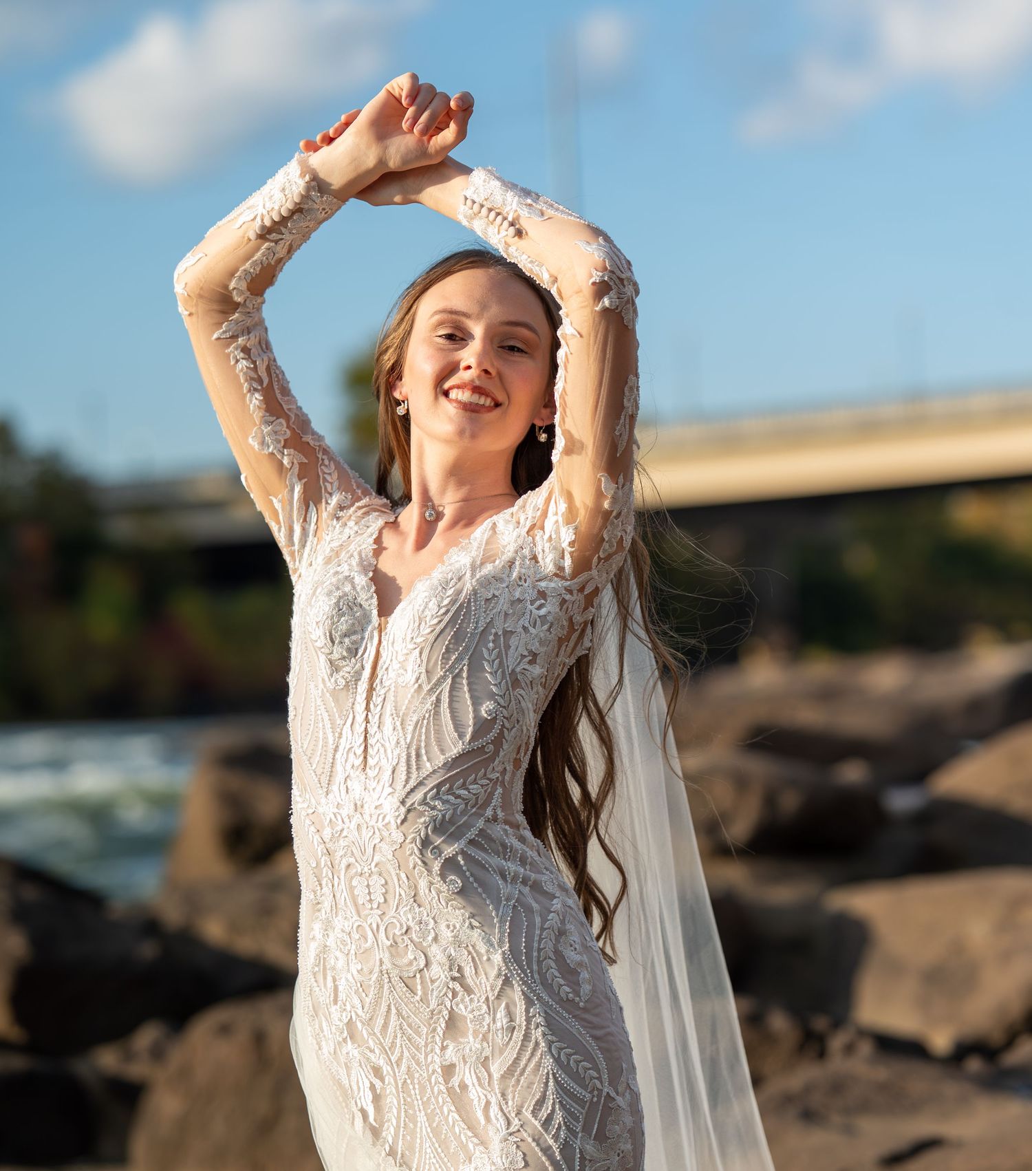 A person in a lace wedding dress poses joyfully at sunset on a rocky beach.