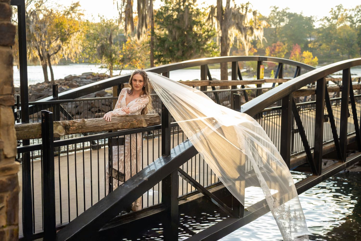 Bride on wooden bridge walkway with flowing veil catching sunlight through trees.
