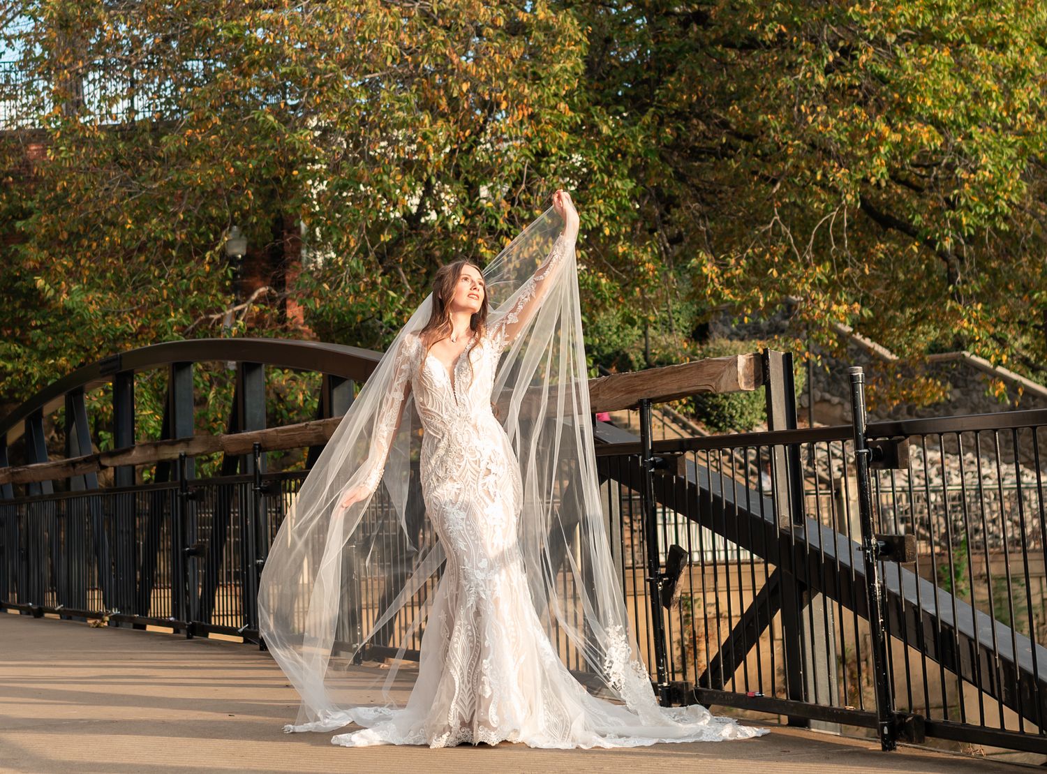 A bride in white lace poses along a railing with autumn trees in the background.