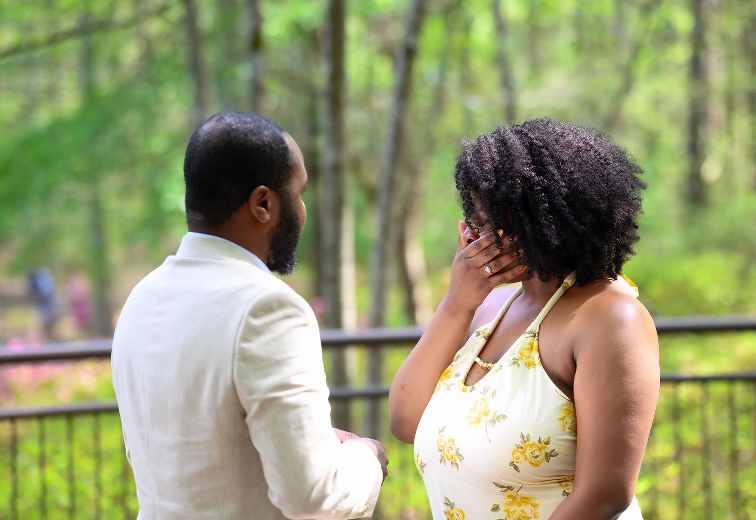 A woman covers her mouth in surprise during an outdoor marriage proposal while a man in a white suit holds her hands.